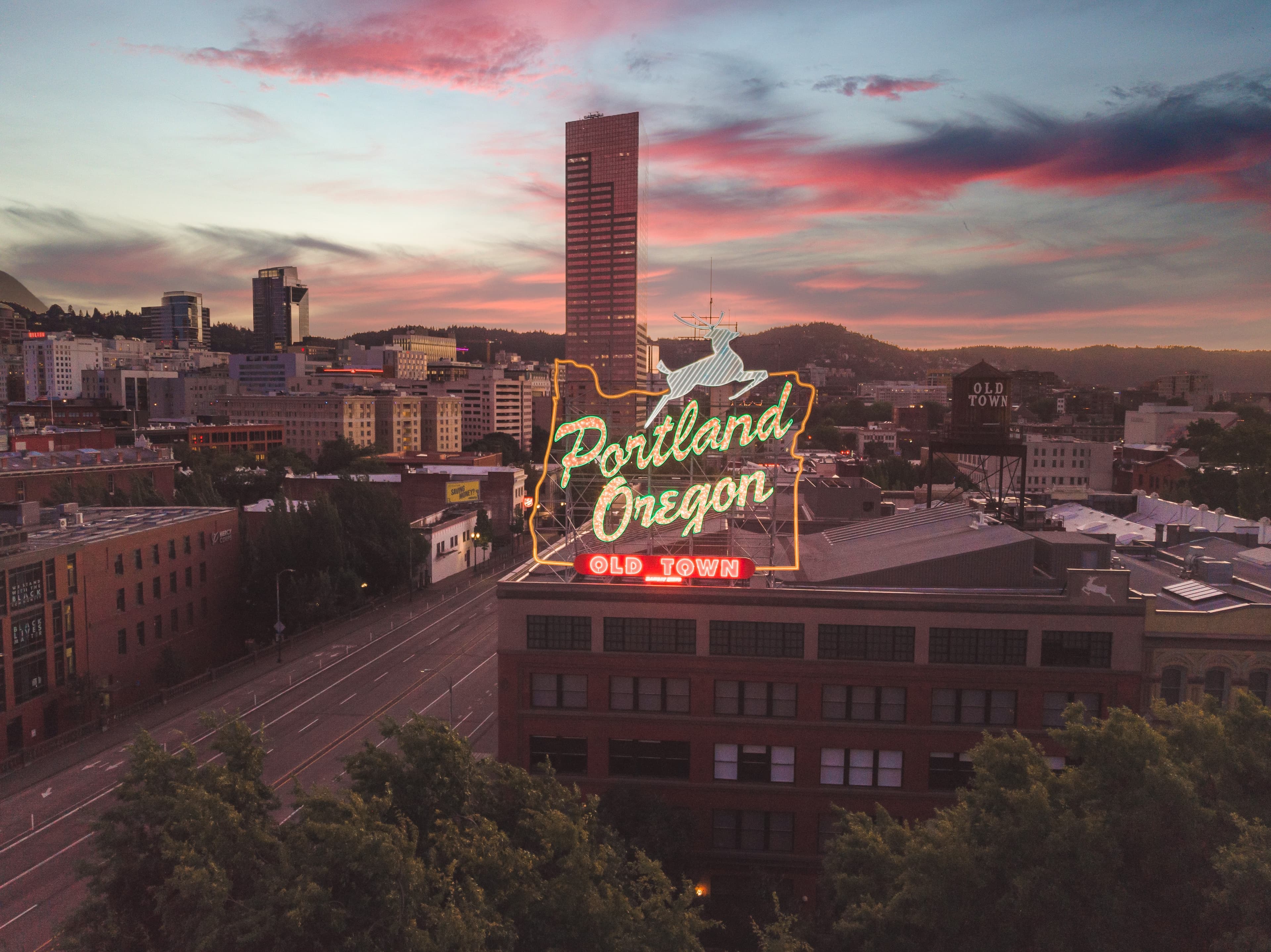 A city view of Portland, Oregon at sunset with pink clouds and a neon sign reading 'Portland Oregon Old Town' with a white deer.