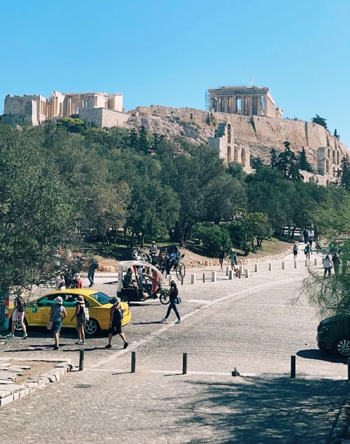 A street view of Athens with people, cars, trees and stone buildings in the distance.