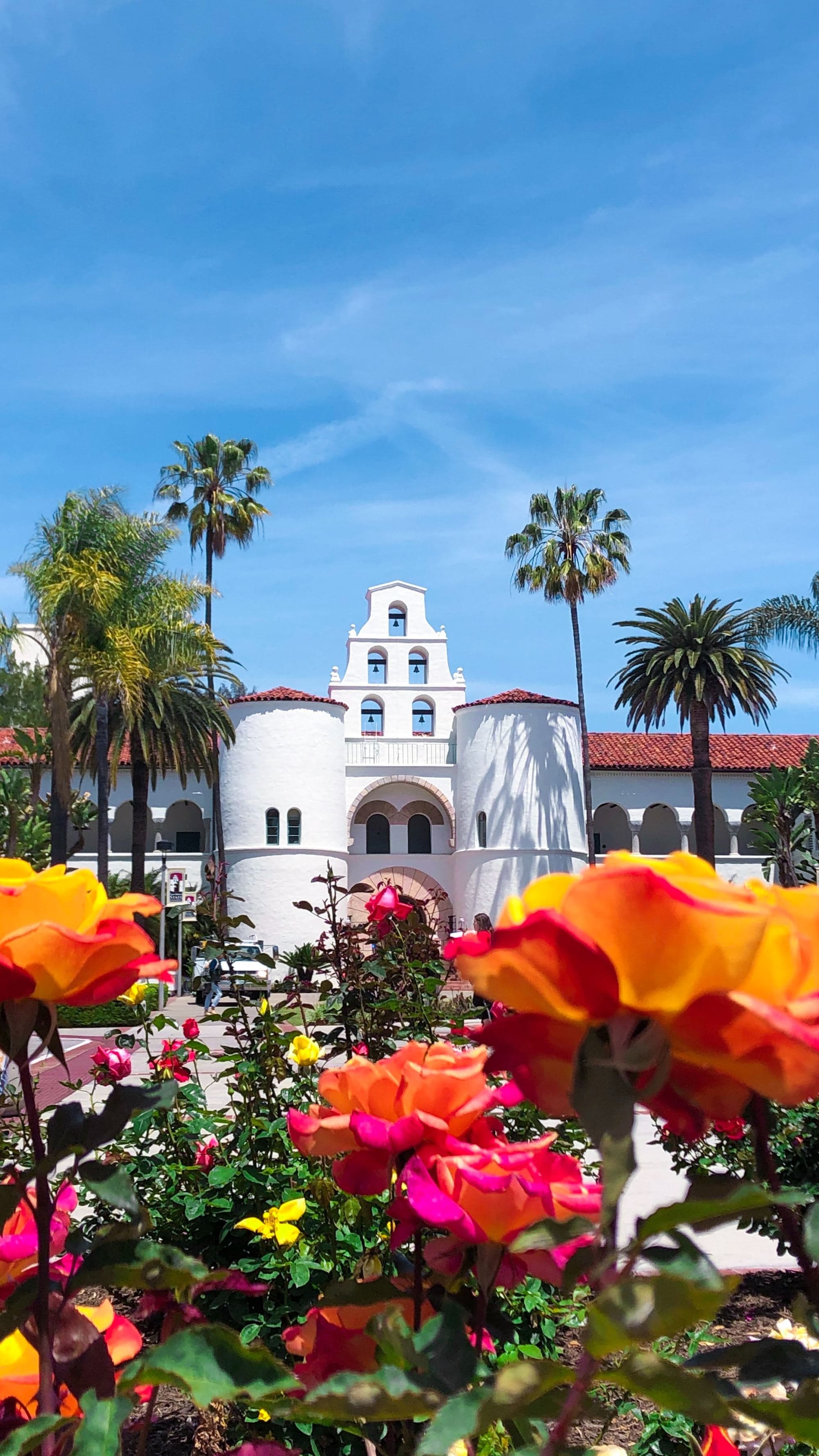 A picture of a white building surrounded with trees and flowers.