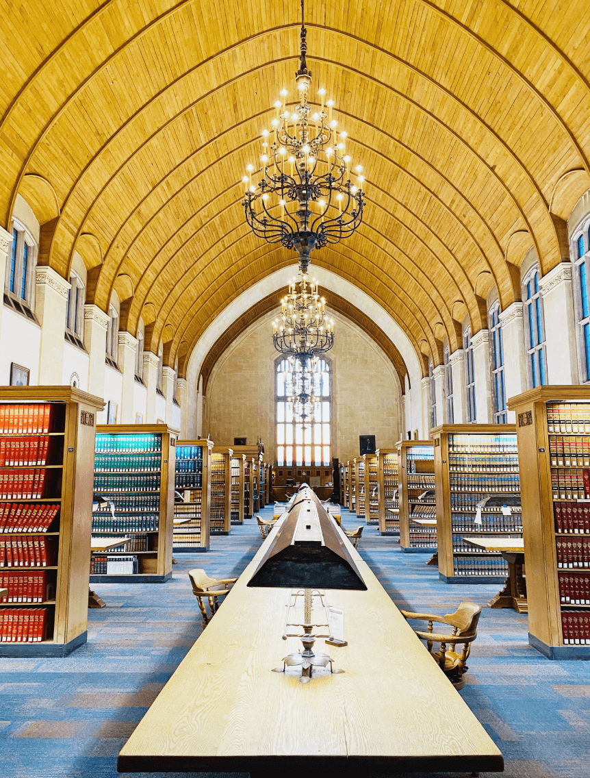 A view of a grand library with arched ceilings, grand chandeliers and one long table down the middle.