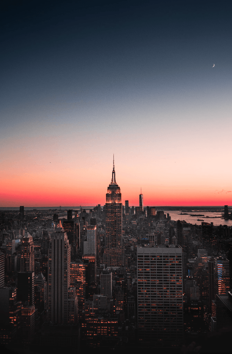 A view of the Empire State Building and New York City skyline at night with a glowing sunset in the background.