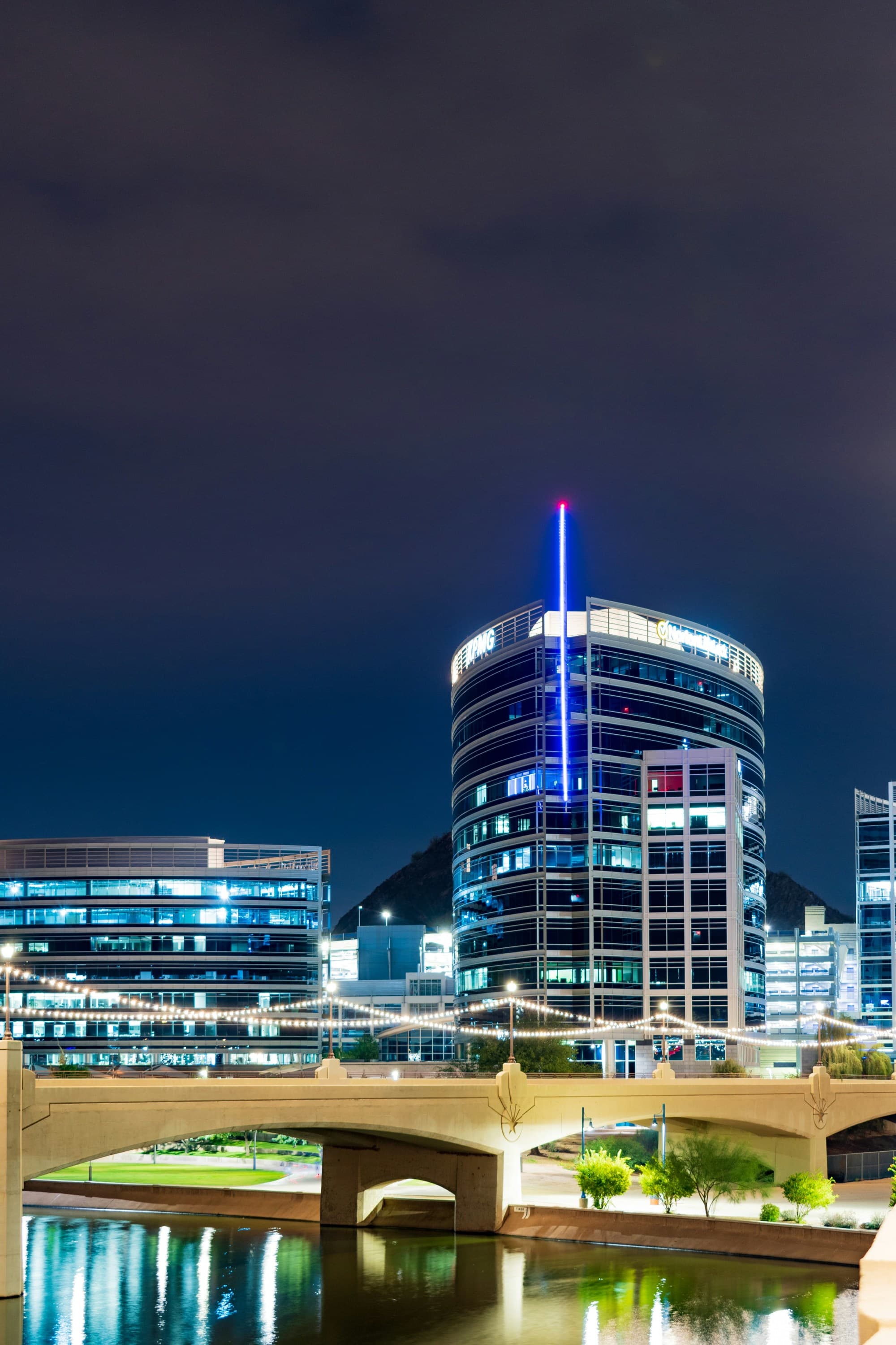 A city skyscraper lit up at night with a large stone bridge over a river next to it.