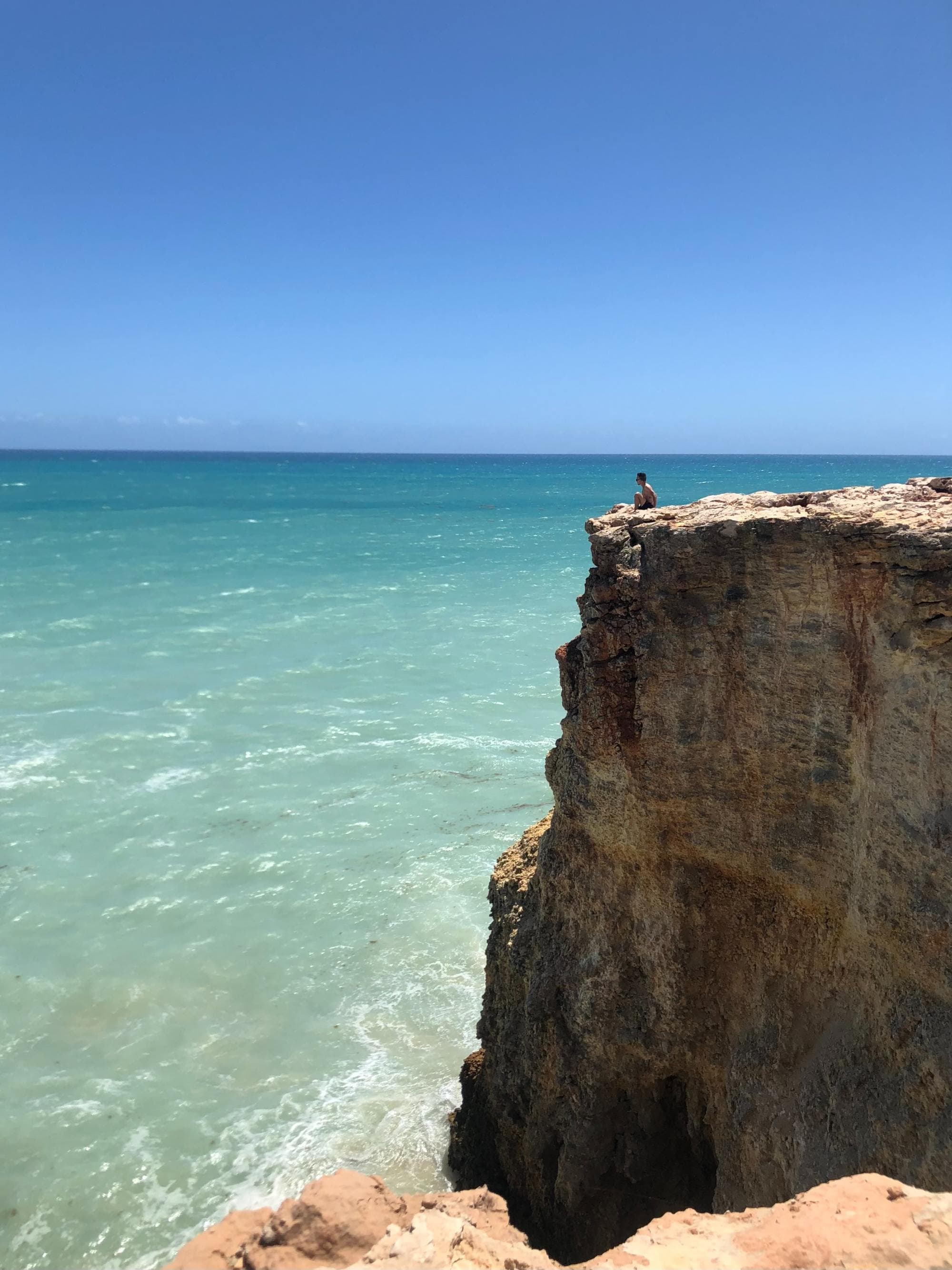 a person sits on the edge of a high stone cliff looking out at the clear light blue ocean on a cloudless day