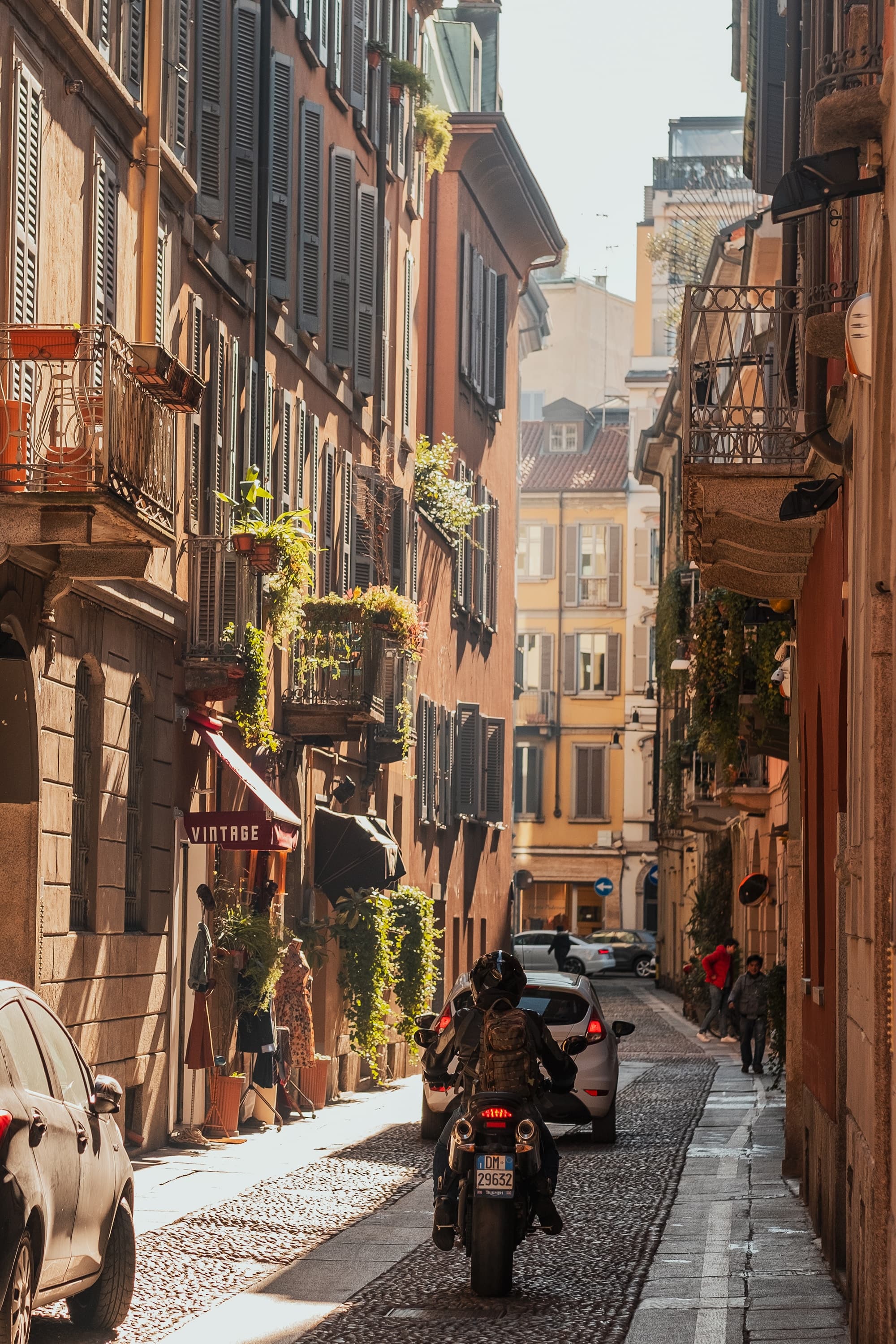 A cobblestone street in Milan with a scooter and some cars.
