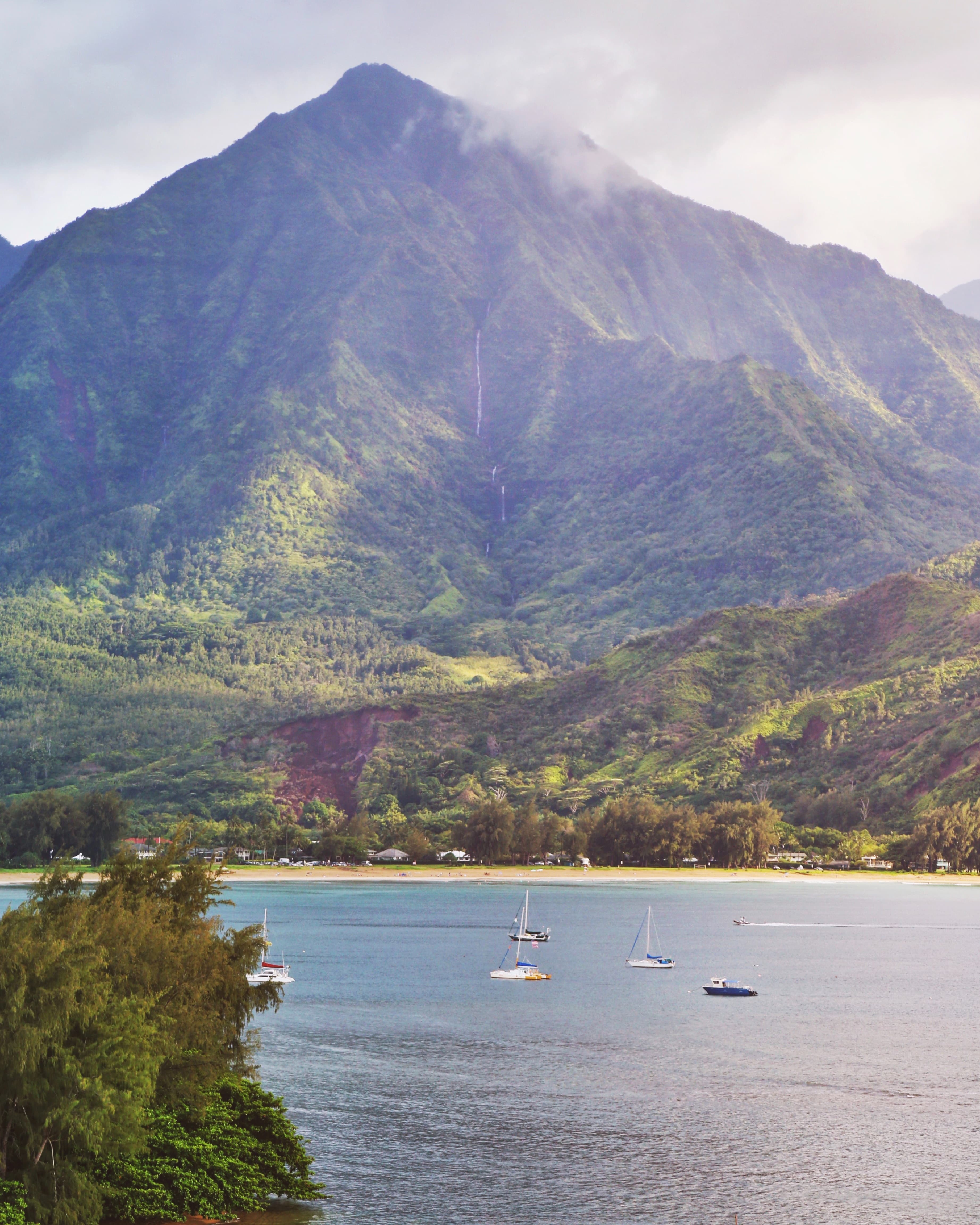 A beautiful beach and sailboats on water embraced by a Hawaiian cove and mountainside.
