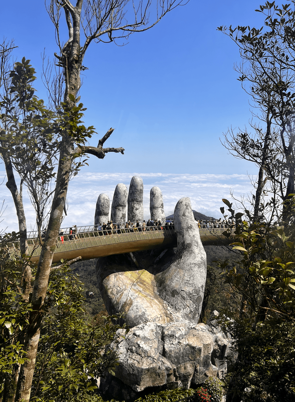 A view of a large stone hand with a bridge built onto it and people walking across. There are also two sets of tall trees and a forest in the surrounding areas.