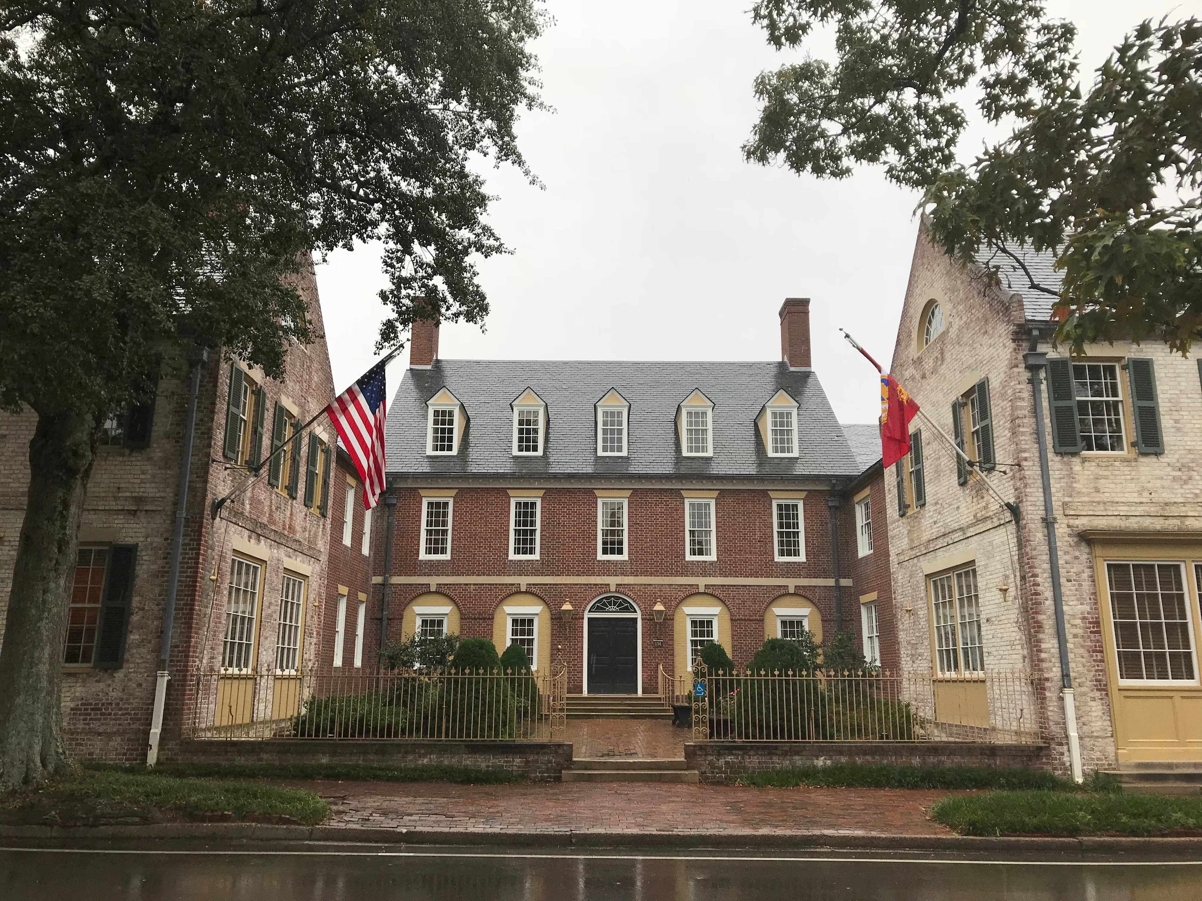 A red brick building behind a gate with white brick buildings, trees, and flags on either side