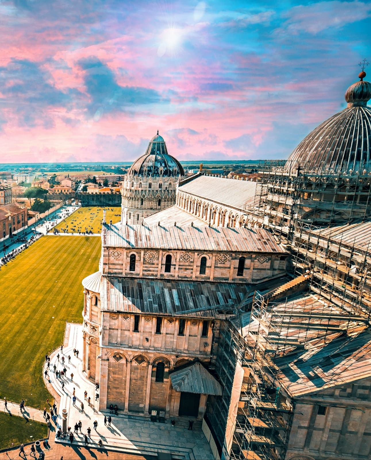 A view of old architecture in Pisa, surrounded by a green lawn beneath a pink and blue sky.