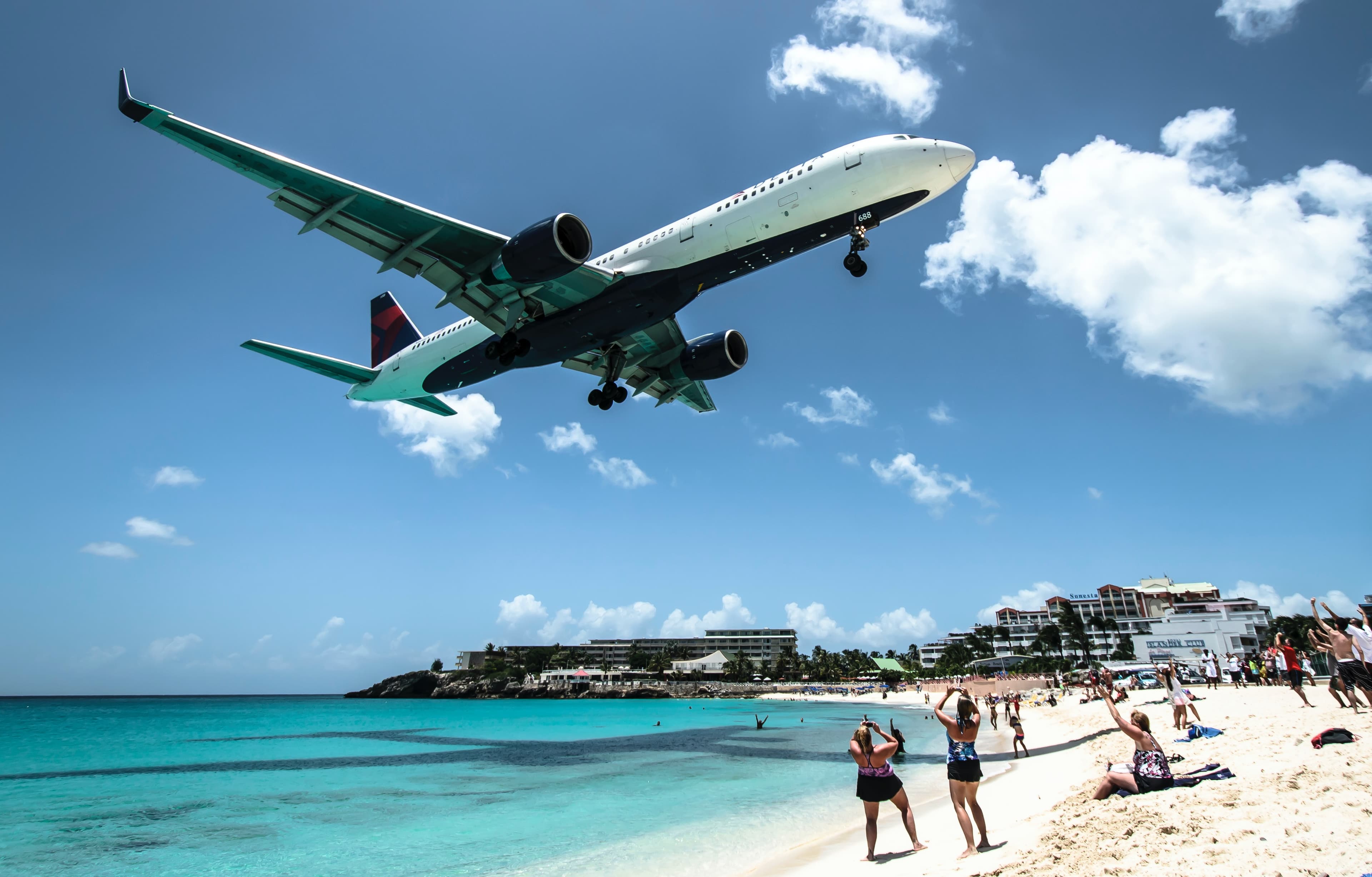 A white and blue passenger plane flying over a blue beach with white sand and tourists in St. Martin.