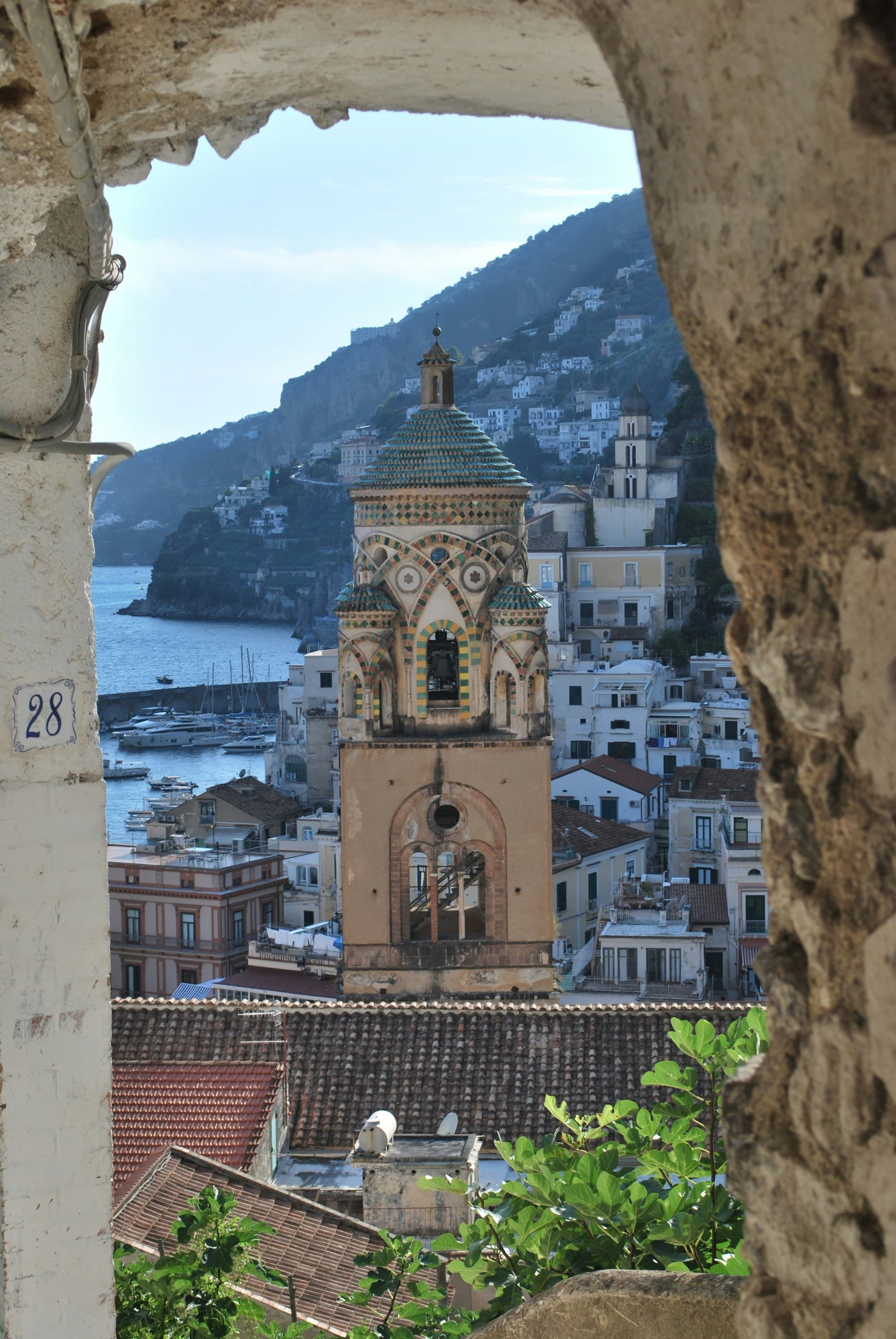 A stone archway highlighting an old bell tower and coastal town in the distance with the sea and mountains showing.