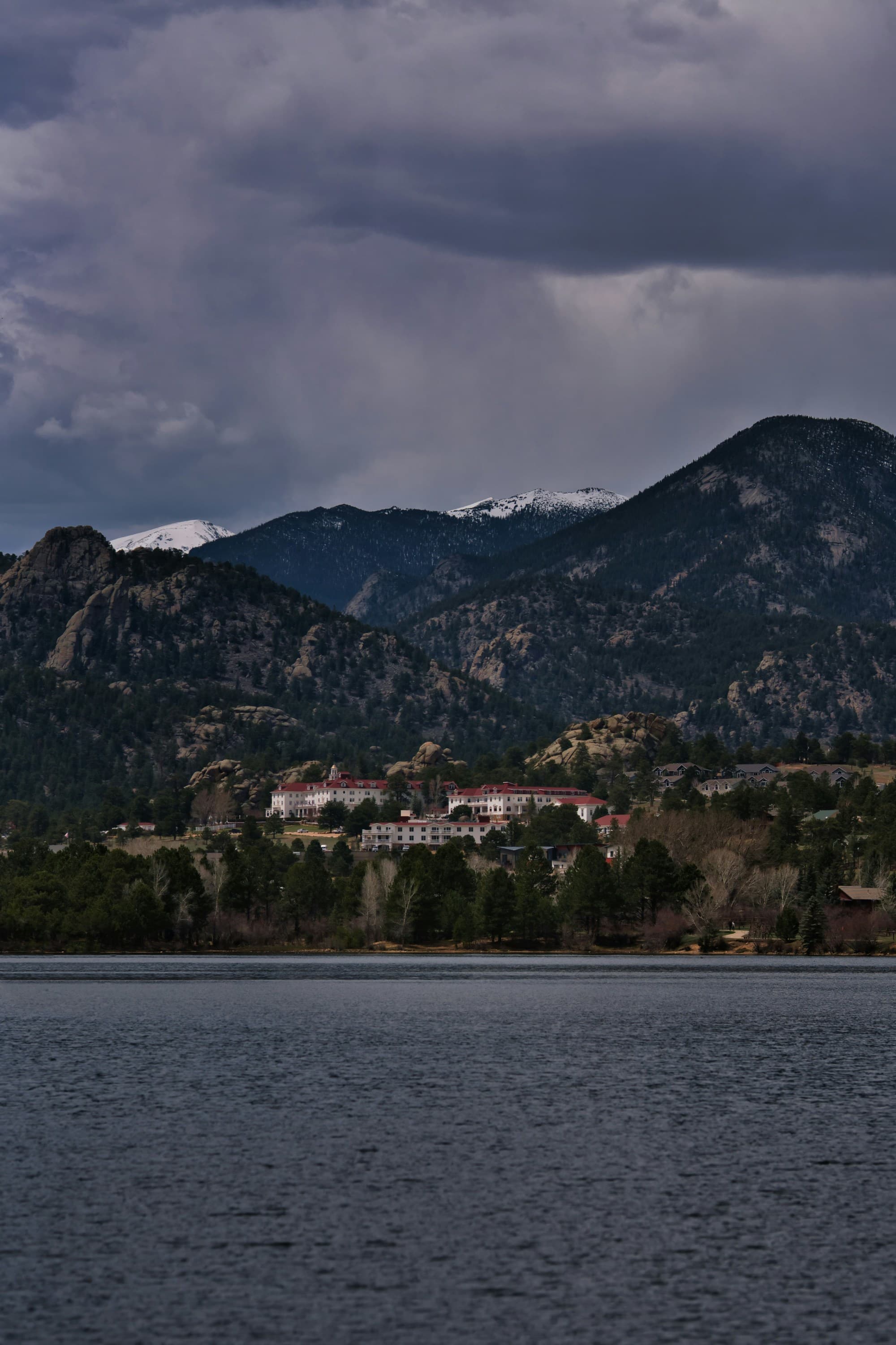 A view of a dark body of water with a white villa nestled into a green mountain under a cloudy grey sky.