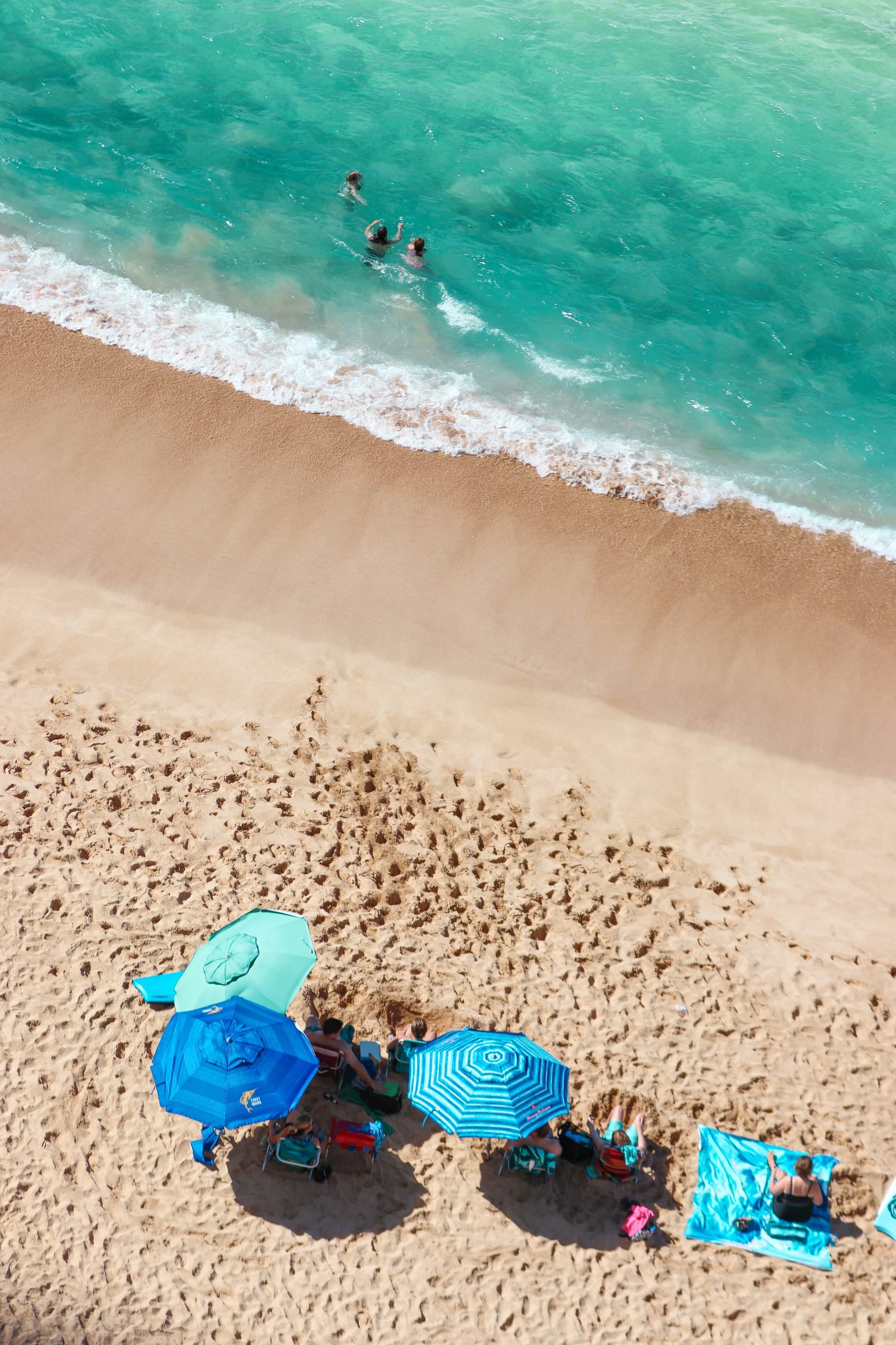 An aerial view of the beach with some of the people lying on the sand under beach umbrellas, while others swimming in the water.