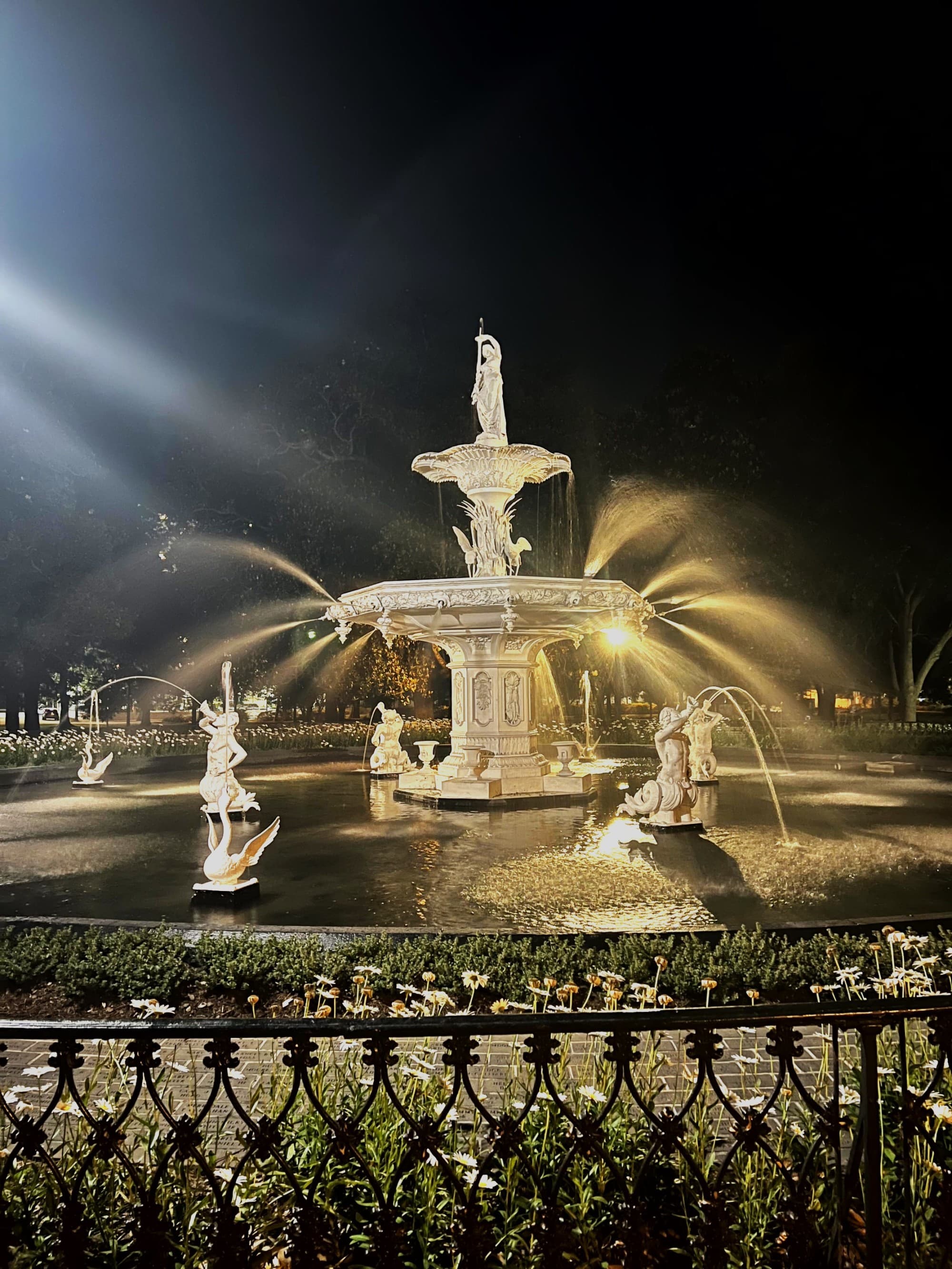 A night view of an ornate, illuminated fountain with statues, set in a dark park setting.