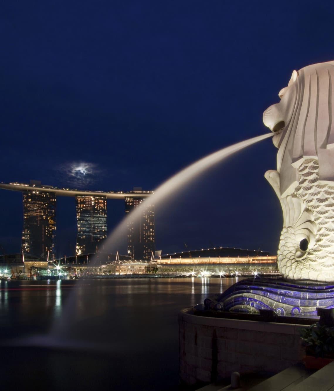 An image of a large white sculpture with water spraying from the mouth into the ocean and three tall skyscrapers illuminated in the background.
