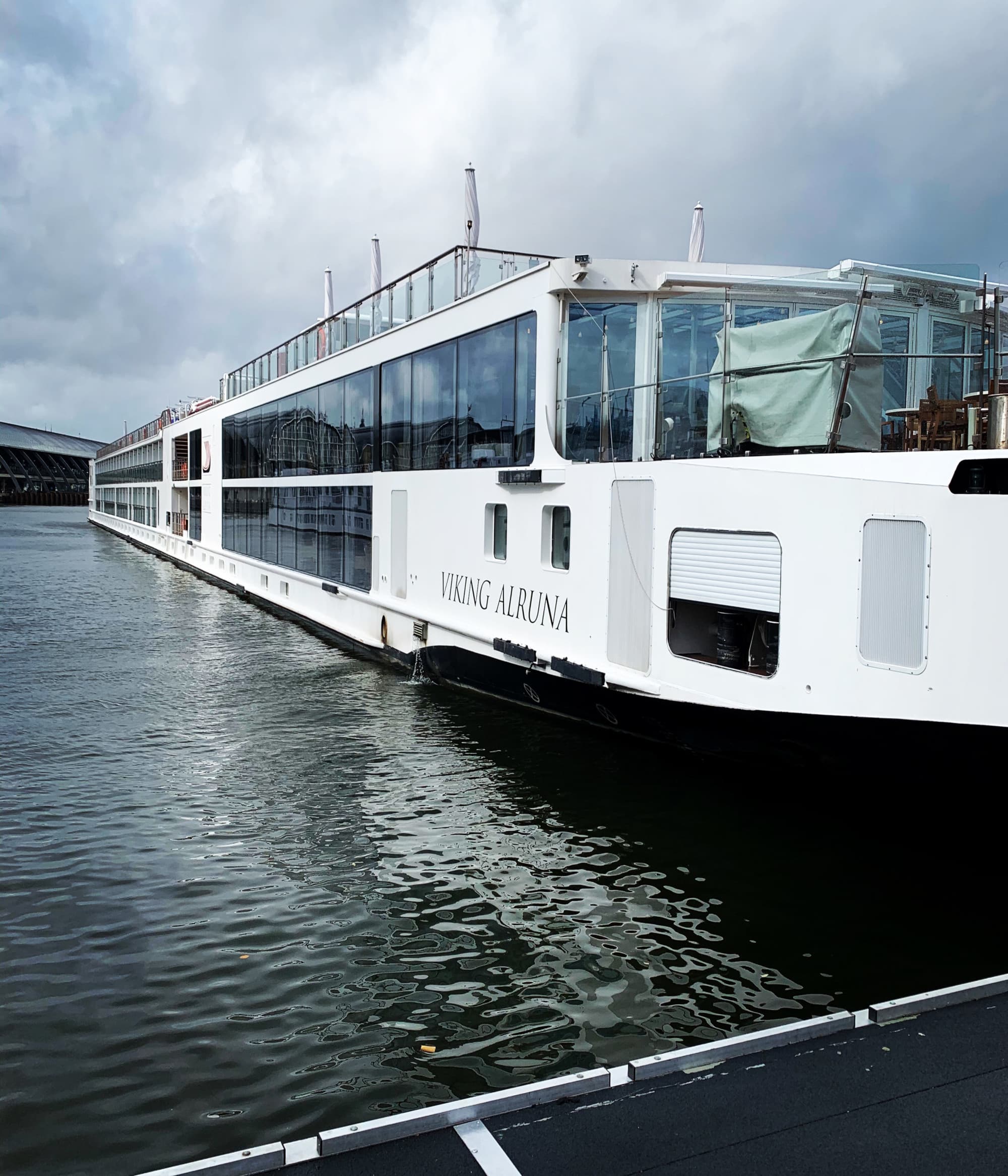 A large white riverboat named ‘Viking Alruna’ is docked on a calm body of water with a bridge in the background under an overcast sky.