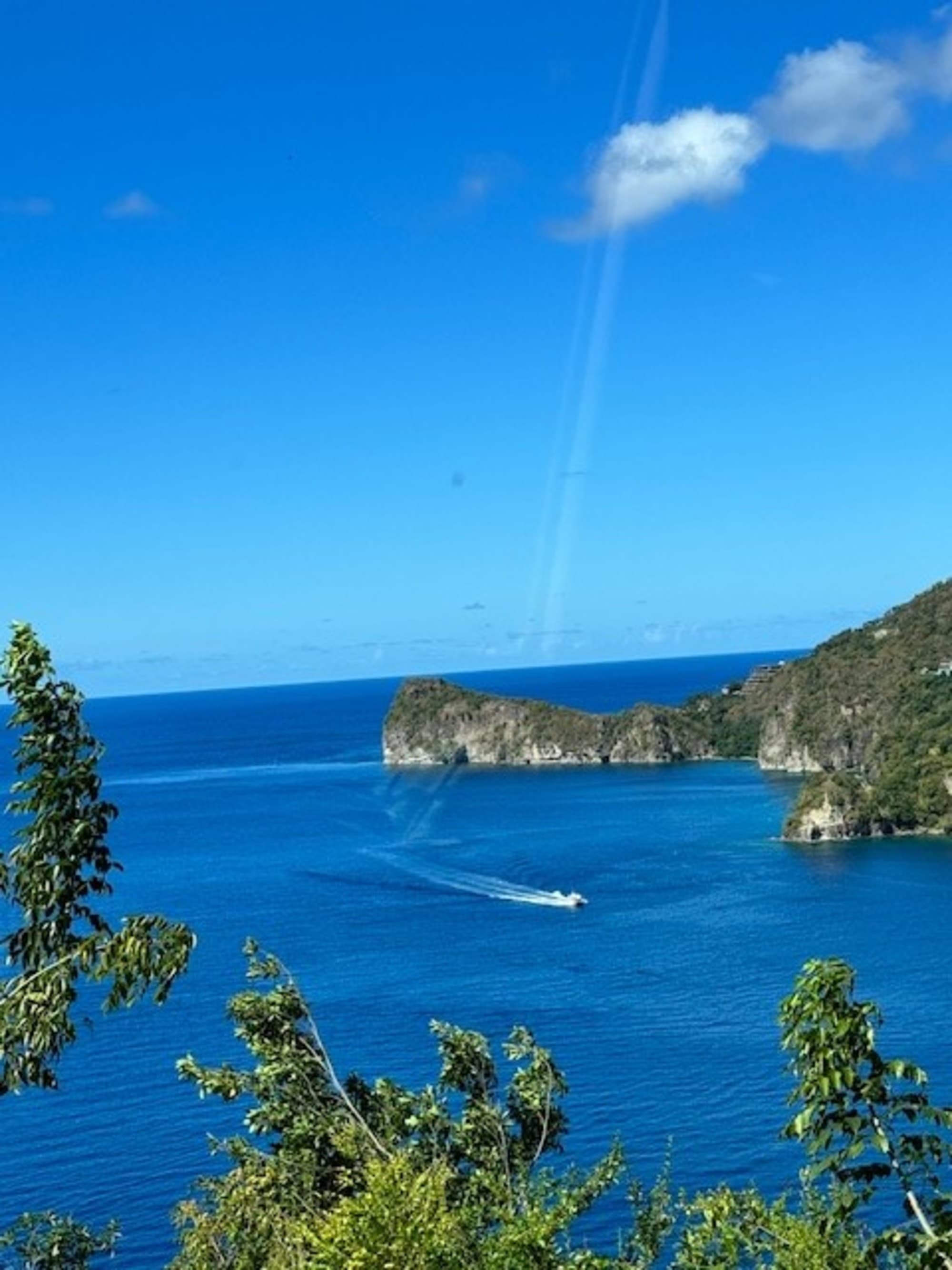 A scenic view of a blue sea with a boat creating a wake, framed by green foliage and cliffs under a clear sky.
