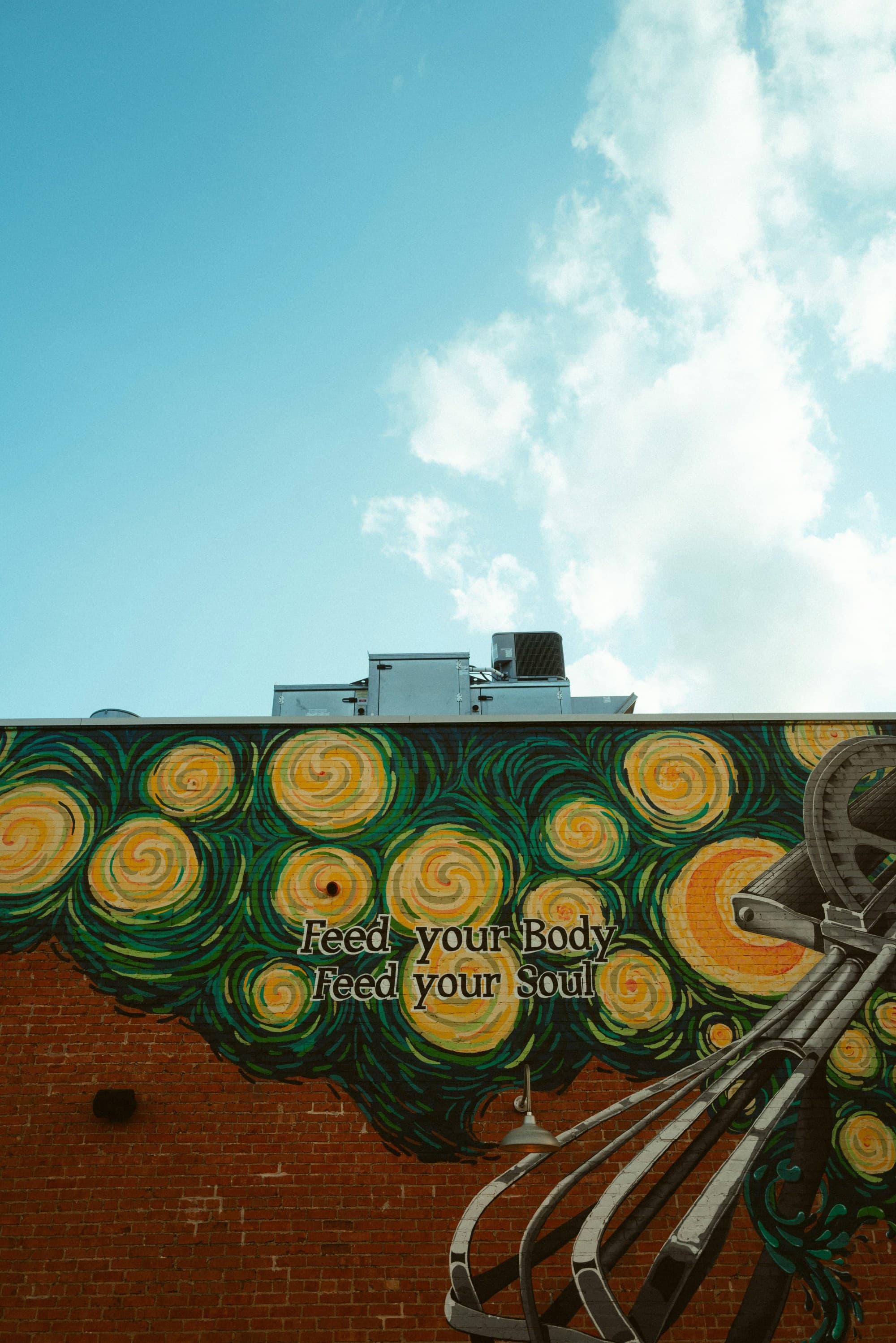 A vibrant mural with swirling patterns reminiscent of Van Gogh’s “Starry Night” adorns a building, with the text “Feed your Body, Feed your Soul” visible.