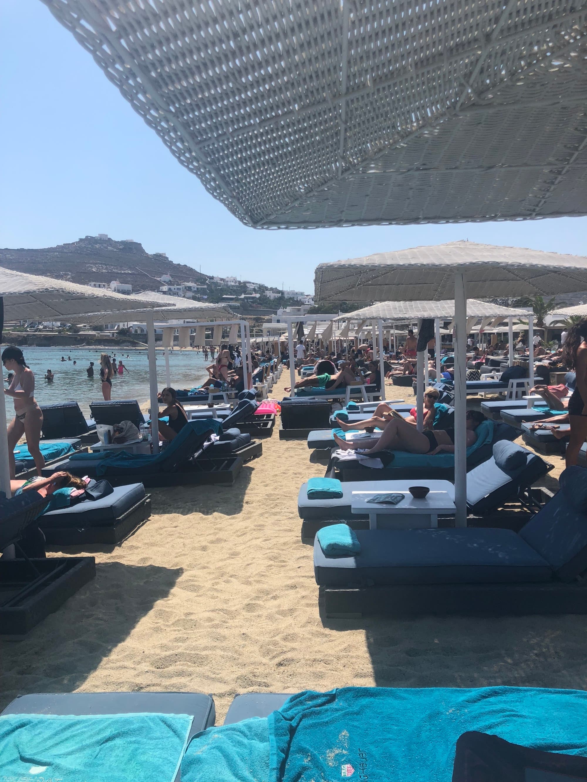 The Kuzina Beach Club in Mykonos has loungers and umbrellas that line the beach in front of the crystal-clear water on a sunny day.