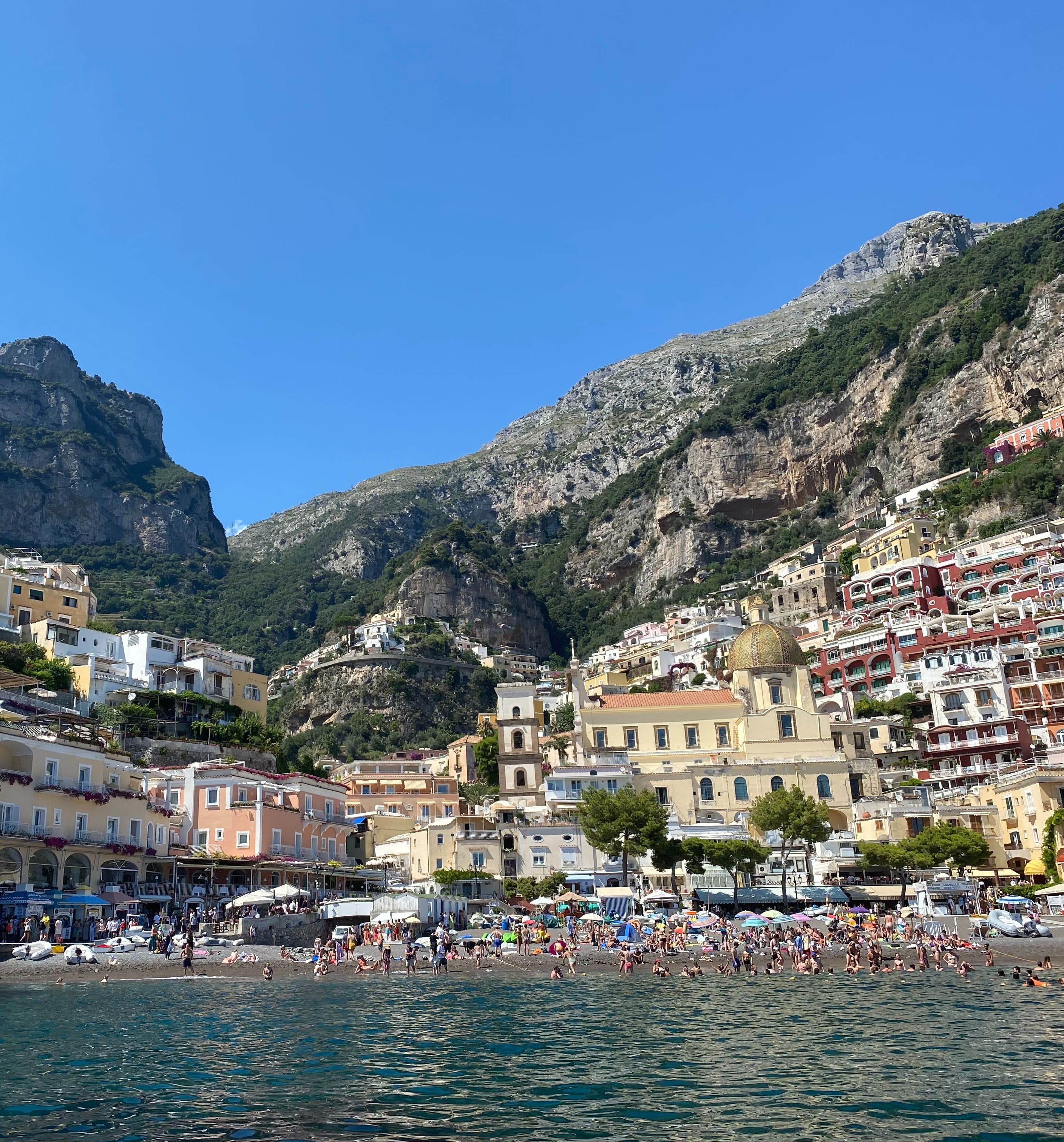 People are gathered, enjoying the beach the city of Positano is nestled into the cliffs behind them on a sunny day.