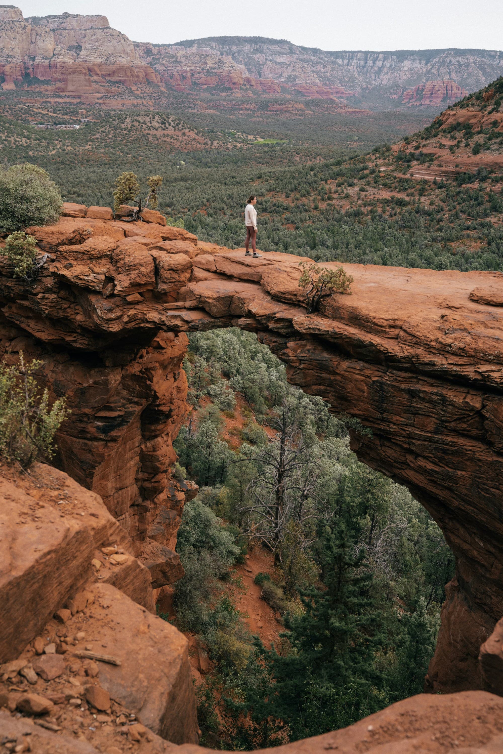 Man in a t-shirt and shorts stands on an arching rock formation overlooking the mountainous region beyond on a sunny day.