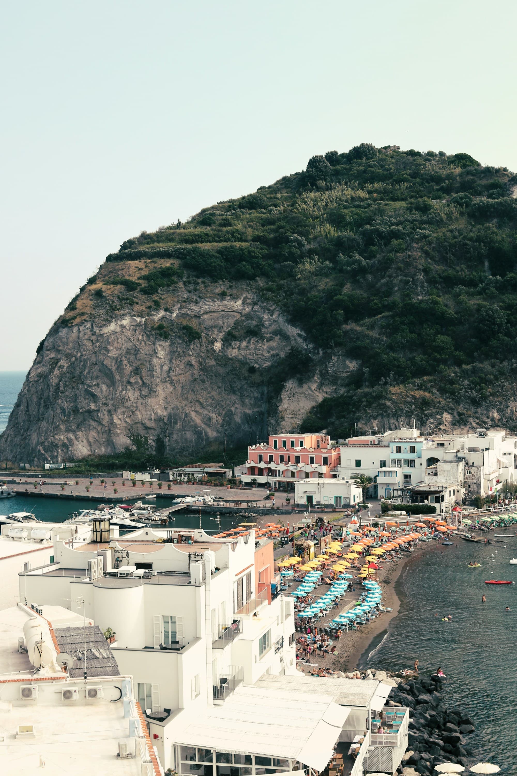The image depicts a coastal scene with colorful beach umbrellas, white buildings, and a large hill or cliff in the background.