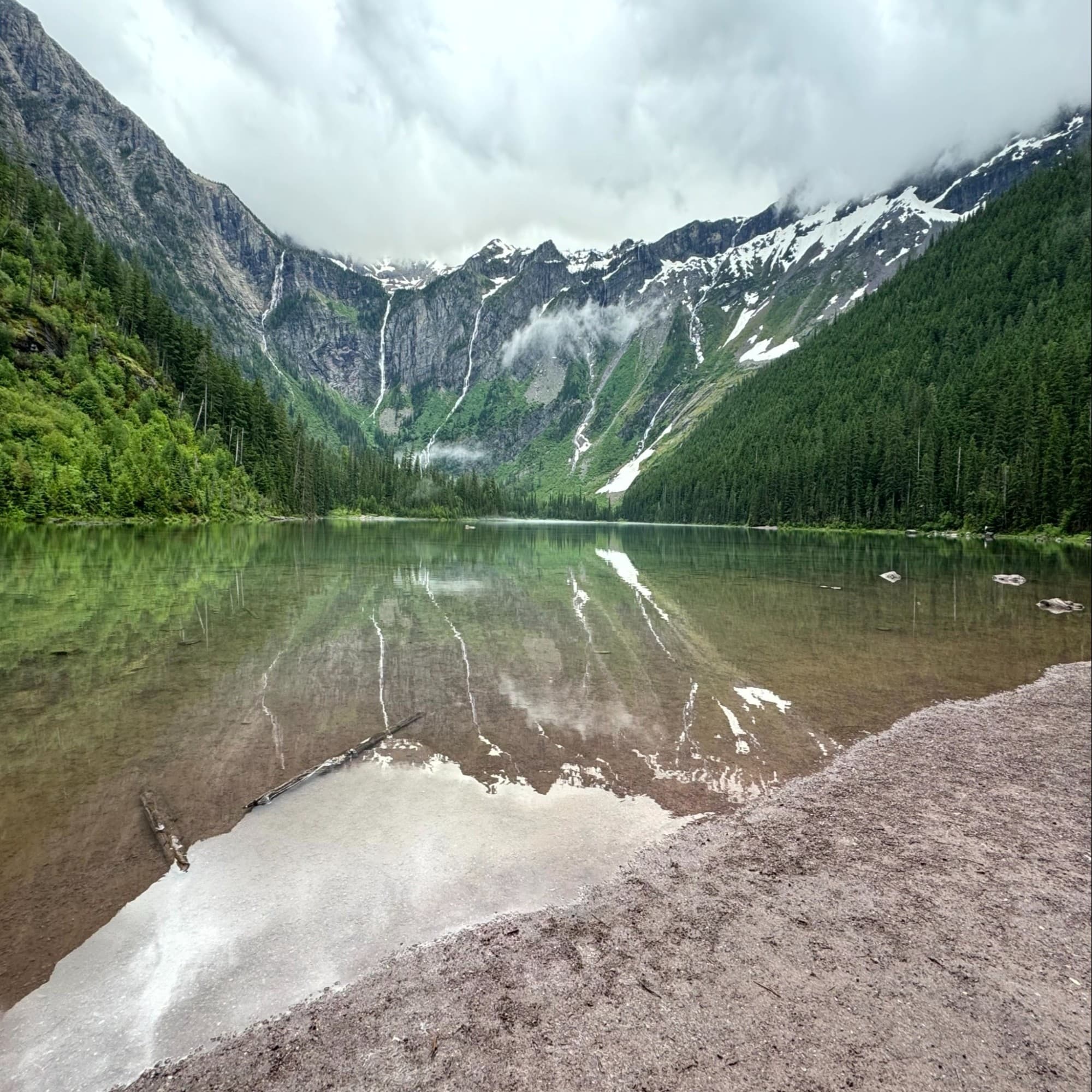 The image captures a serene mountain lake with reflections of snow-capped peaks and forested slopes.