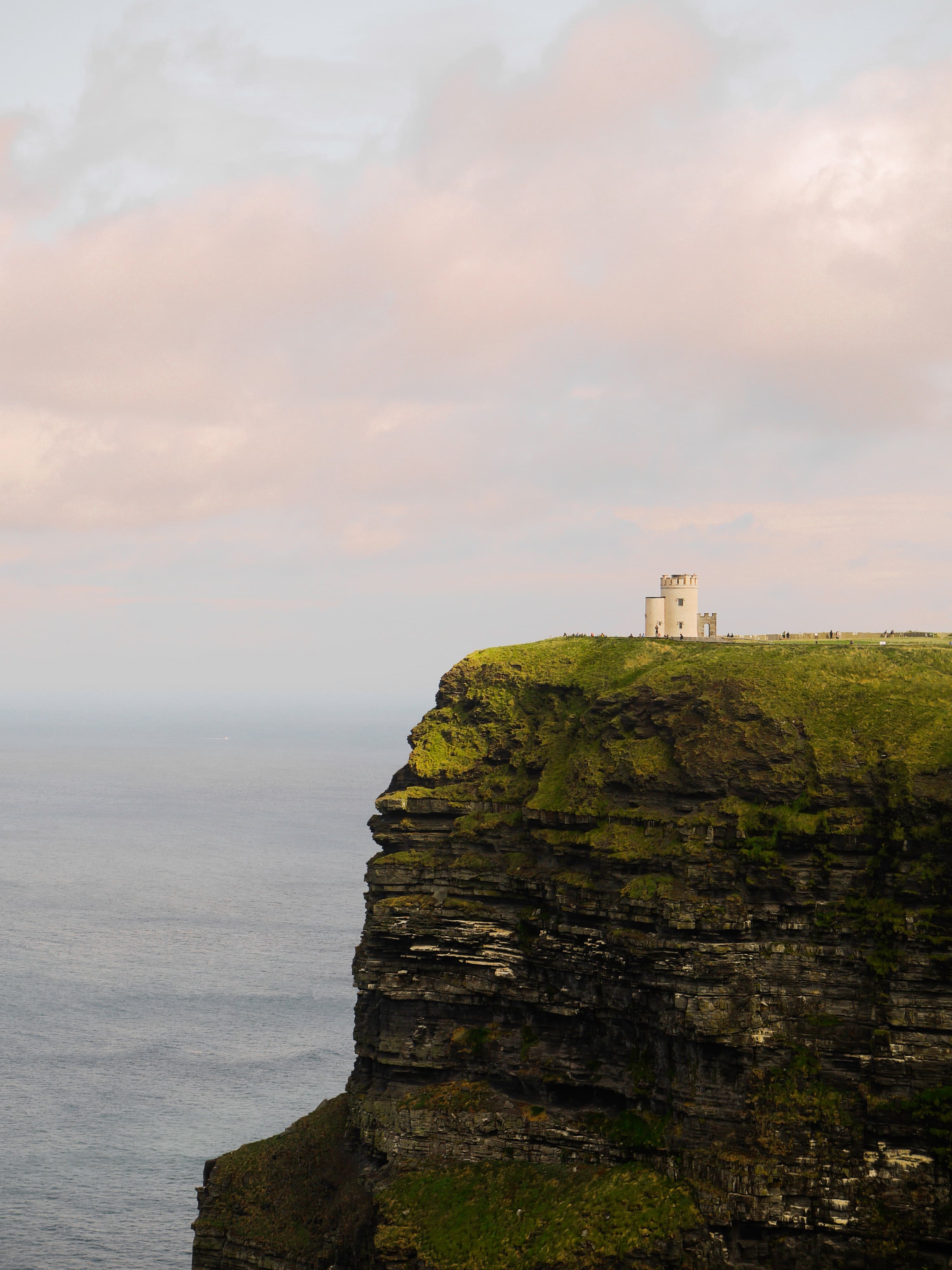 Green cliff with white building next to body of water with pink clouds in the sky during daytime