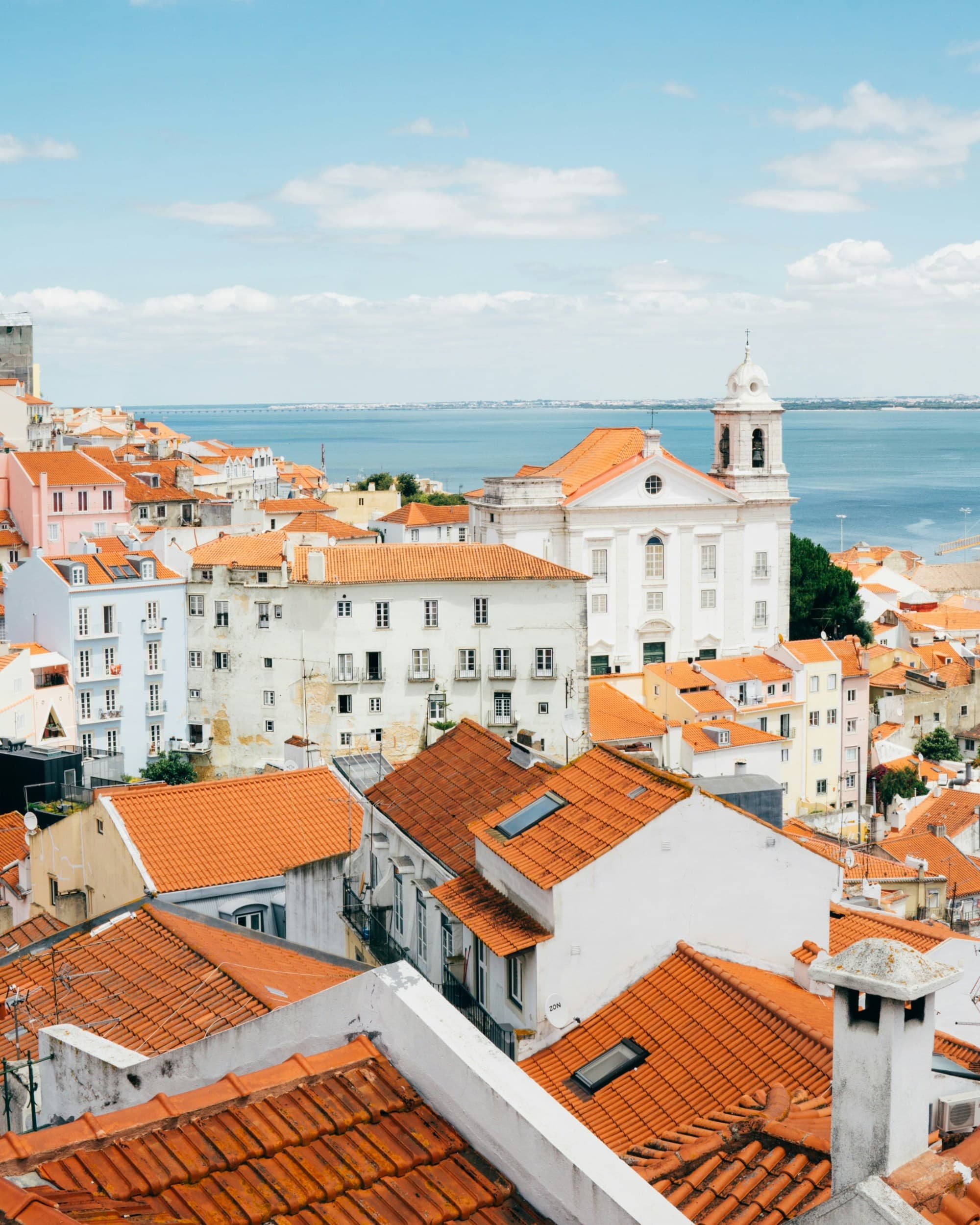 A picturesque coastal cityscape with terracotta rooftops and a church, set against a clear sky, with water in the background.