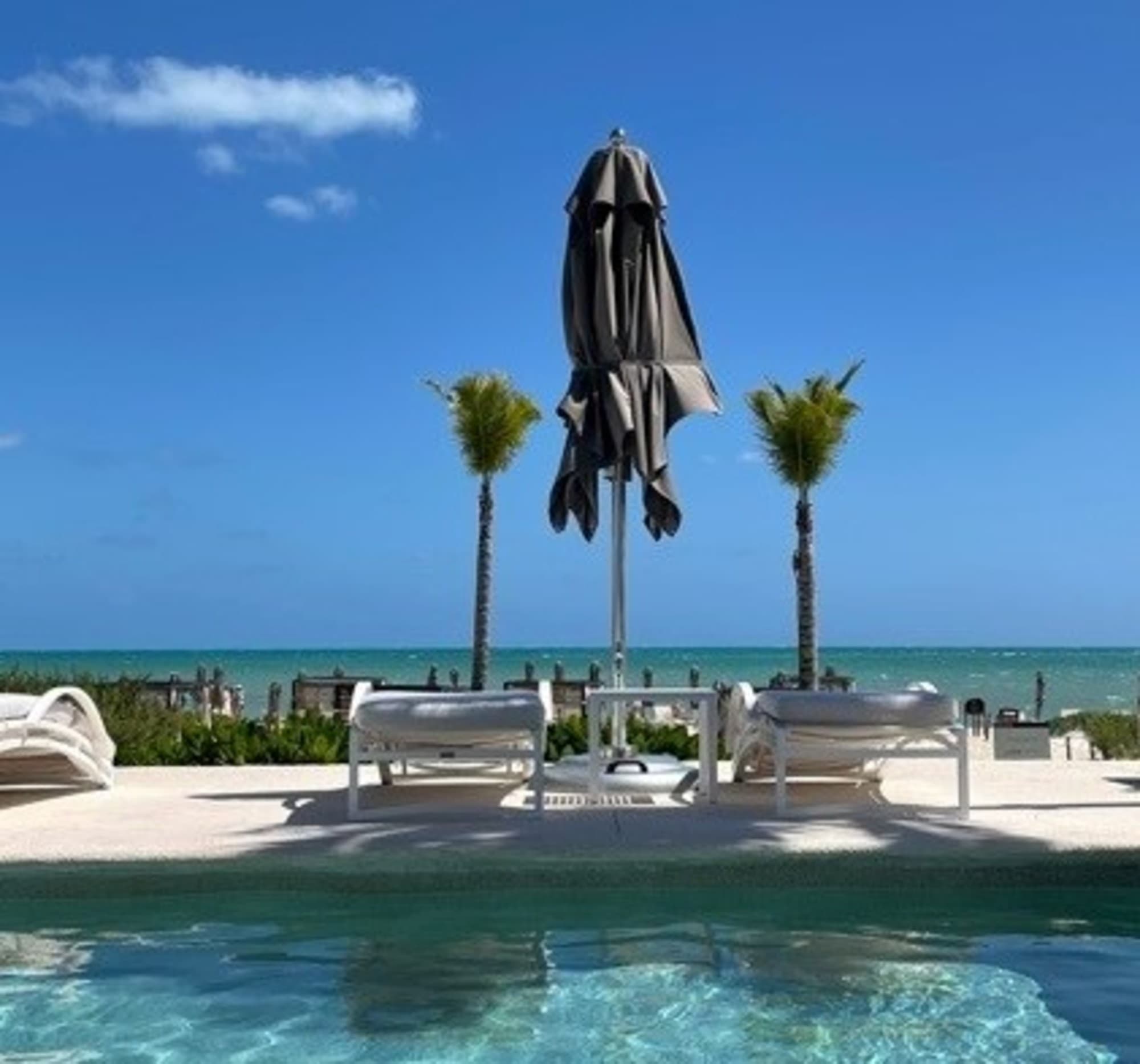 A tranquil poolside view with sun loungers, a closed umbrella and palm trees against a backdrop of a clear blue sky and ocean.