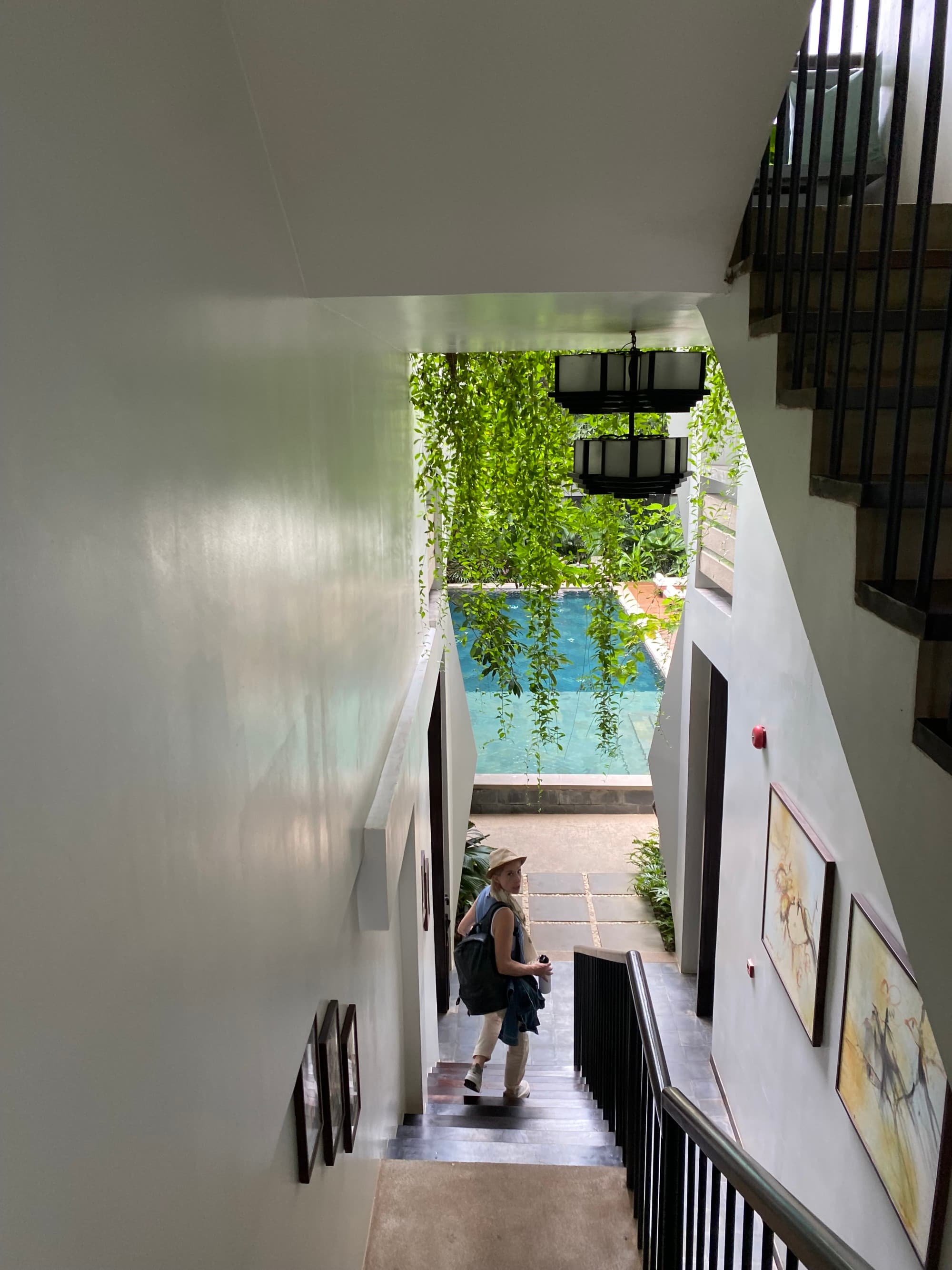A woman standing at the foot of a staircase with large widows and an outdoor pool in the distance surrounded by foliage during the daytime.