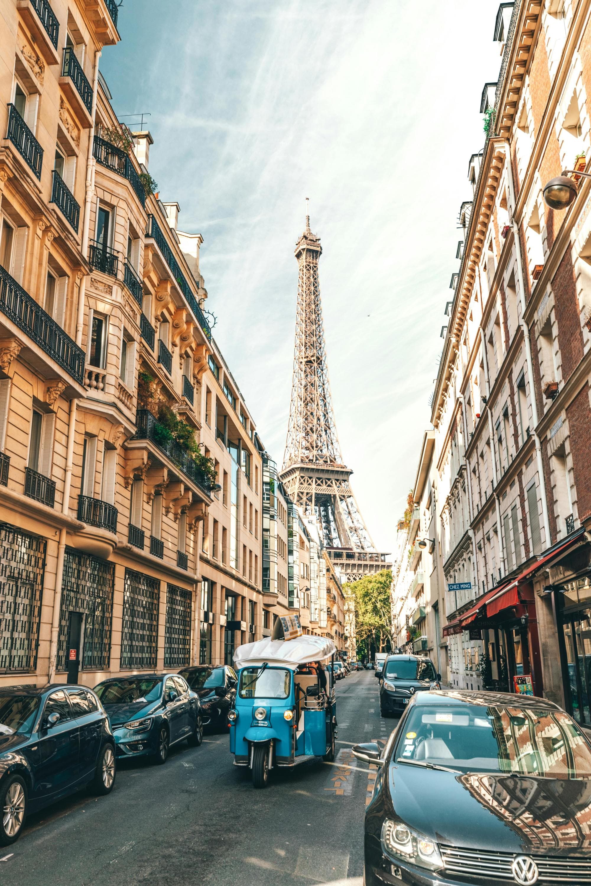 A street view leading towards the Eiffel Tower, with parked vehicles and a distinctive blue three-wheeled vehicle in the foreground.