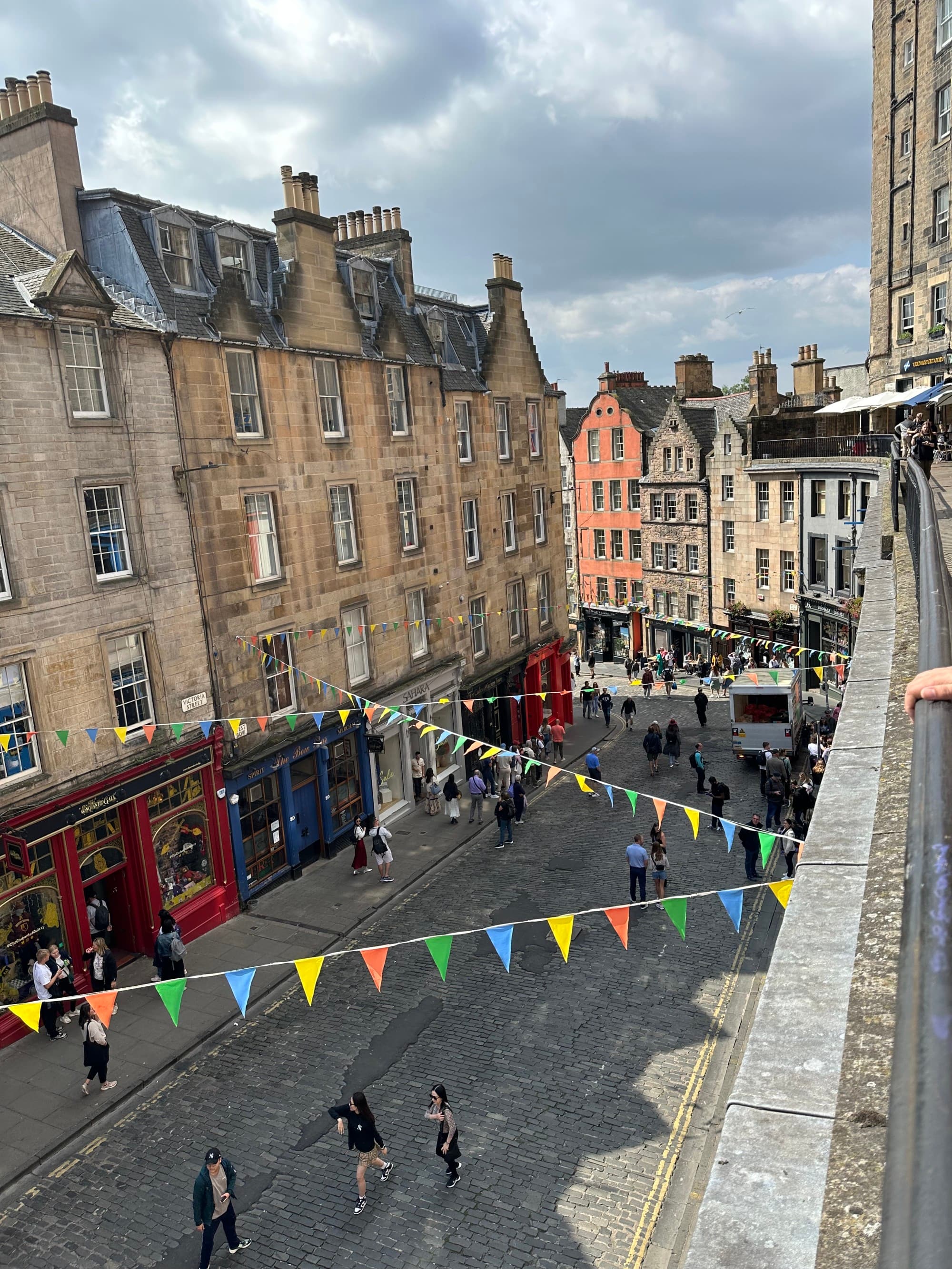 The image depicts a lively street scene with people, colorful decorations, and historic architecture under a cloudy sky.