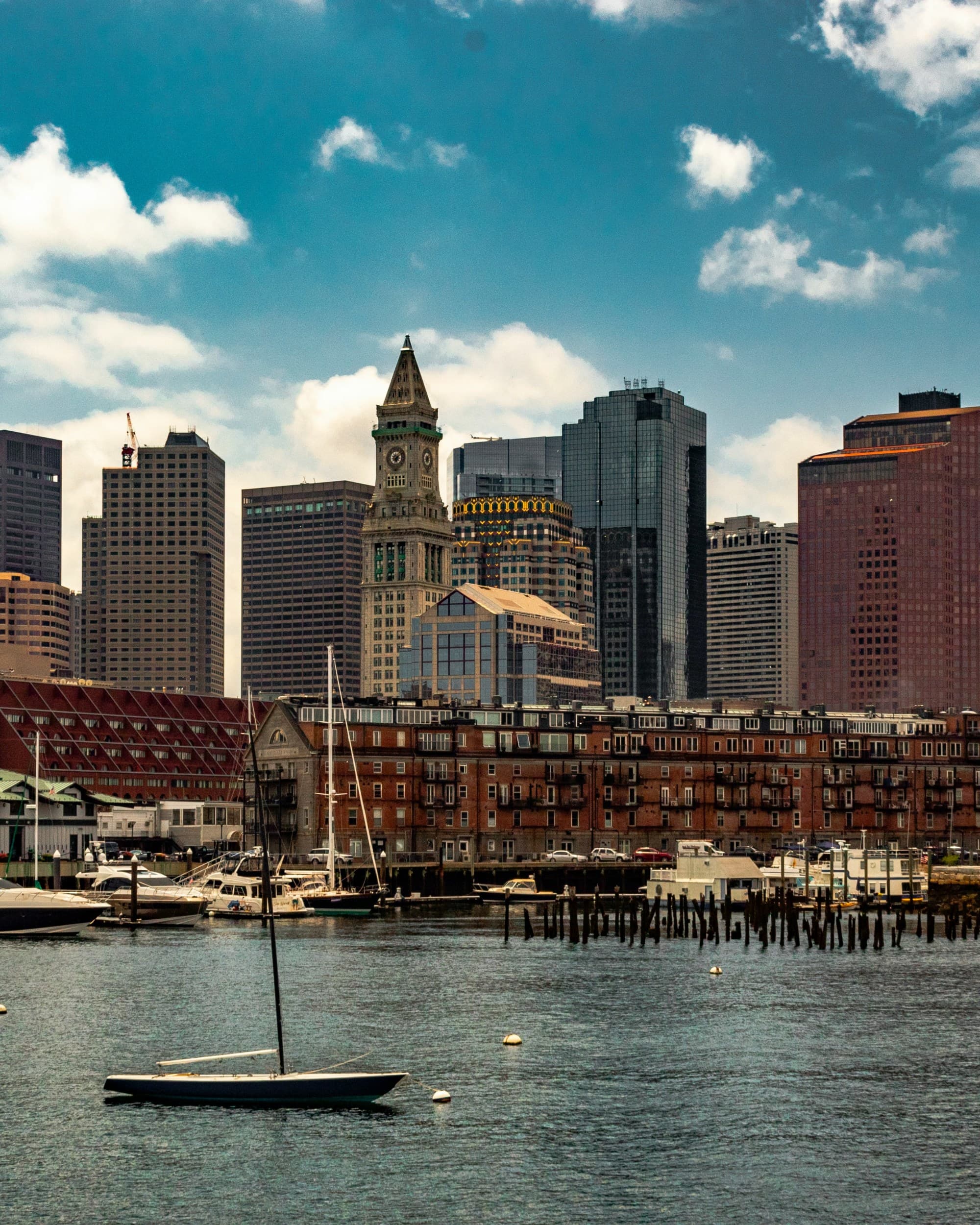 A city skyline with diverse buildings and a sailboat on the water in the foreground.
