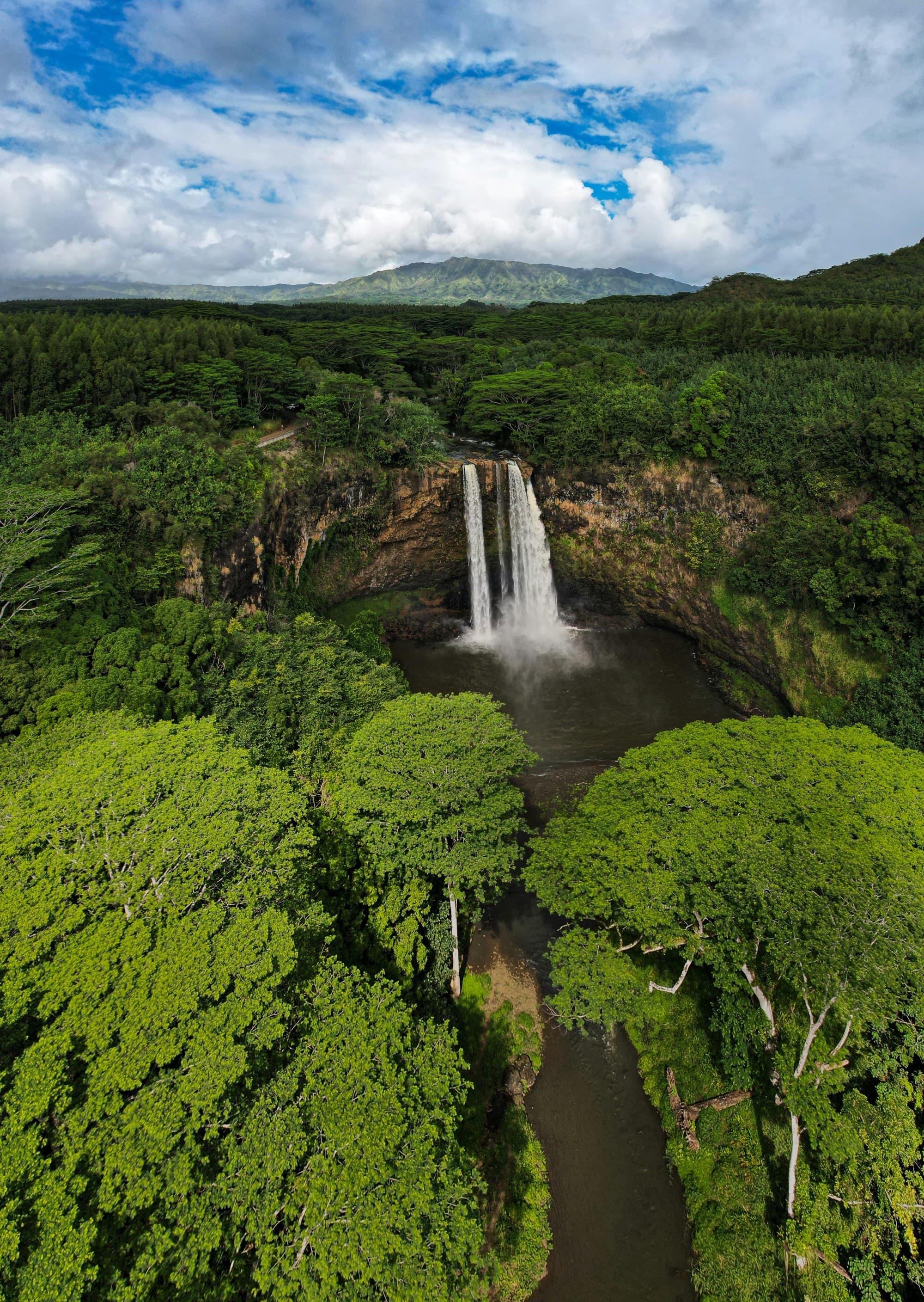 A waterfall, lush greenery and mountains under a partly cloudy sky.