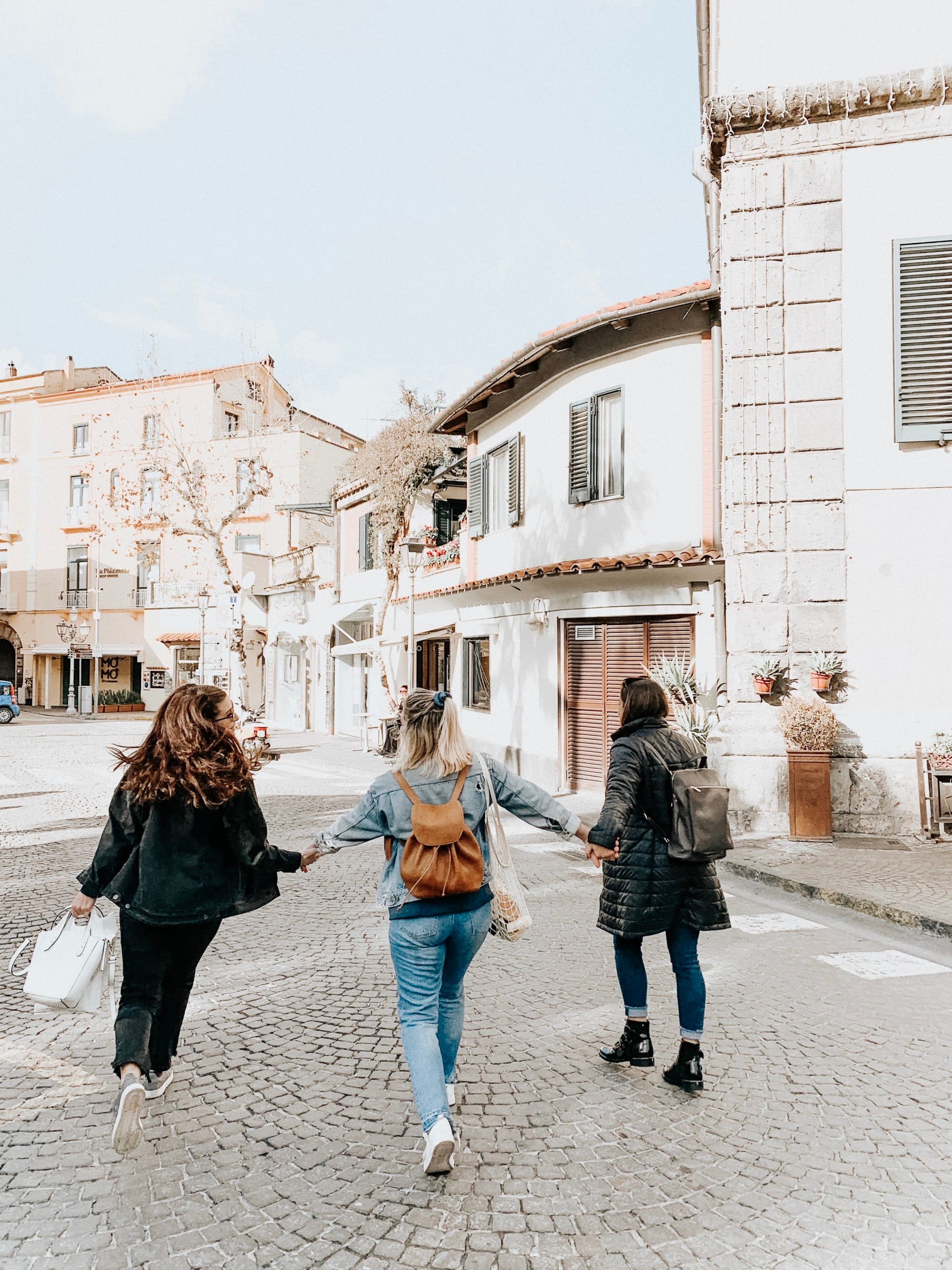 A picture of three girls holding hands and walking on the street surrounded by white buildings.