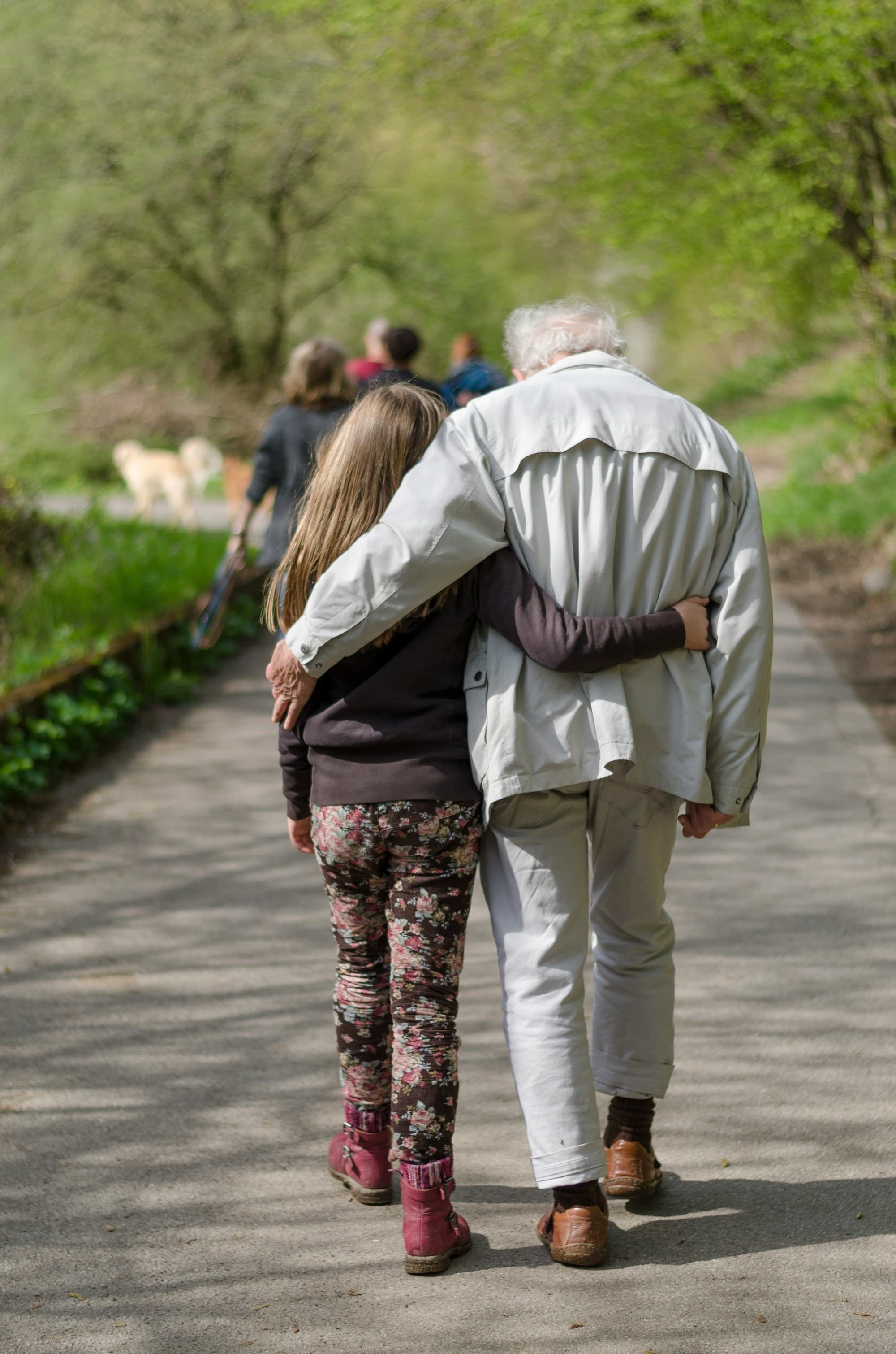 Two companions walk arm in arm along a path surrounded by lush greenery.