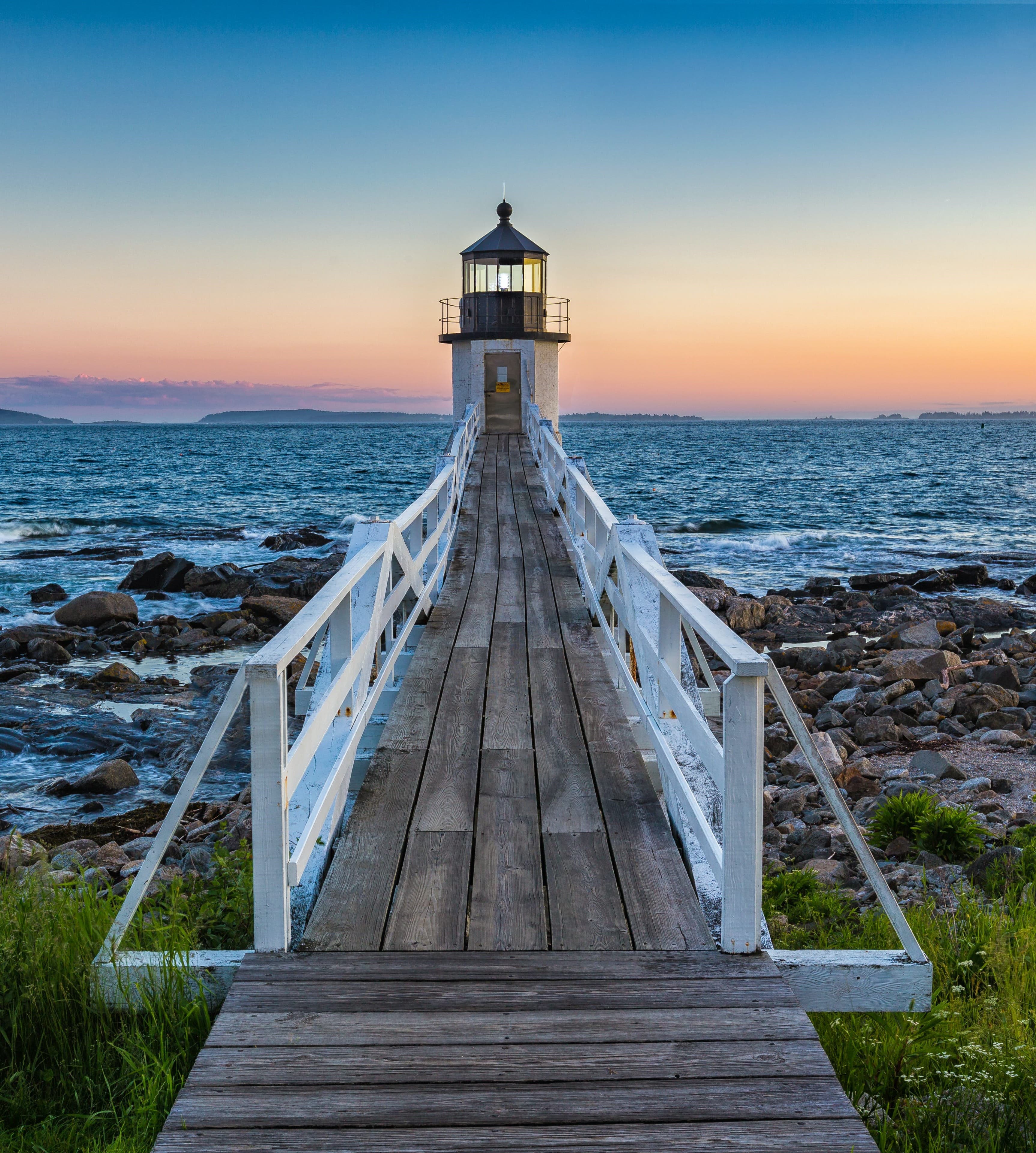 A beautiful wooden bridge with white railings leading to a lighthouse looking over the blue water and light orange sunset.