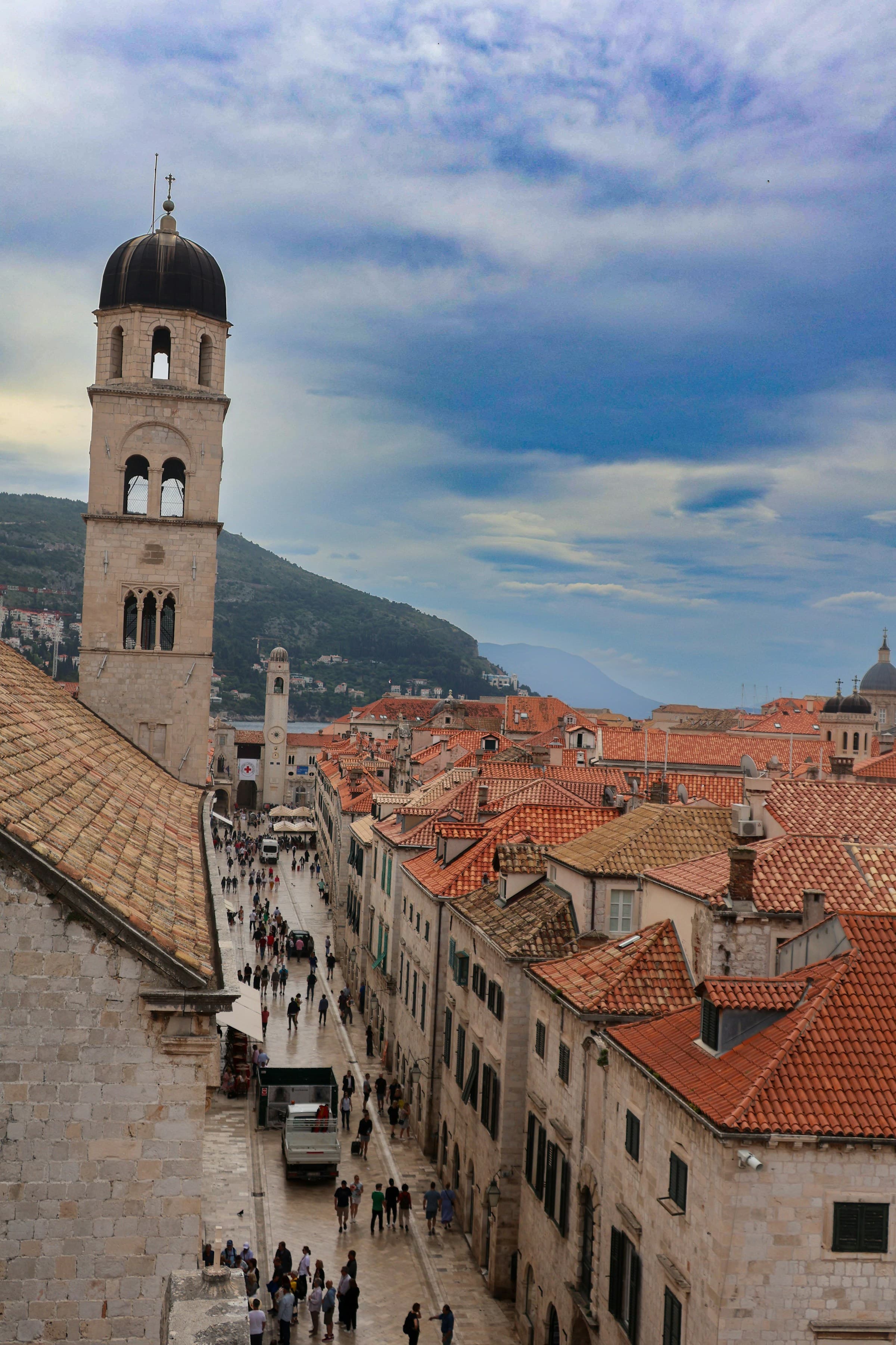 A view of Dubrovnik's Old Town from above, with a bell tower, an alleyway with pedestrians and mountains and sea in the background.