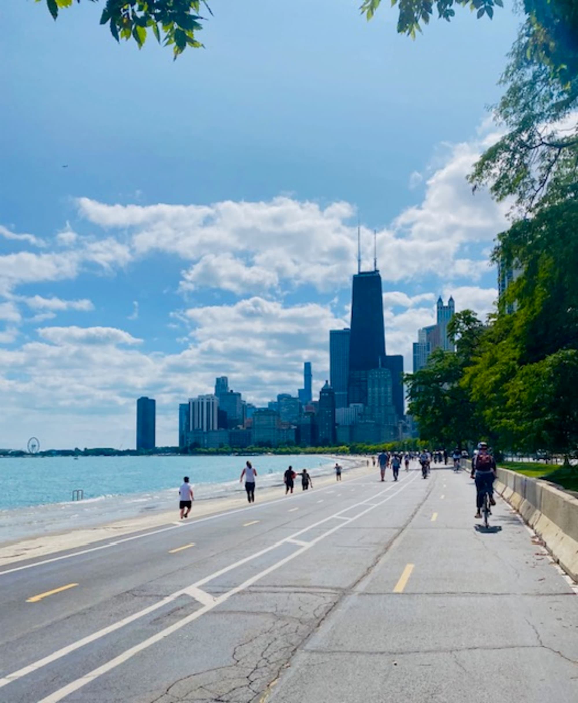 The image offers a picturesque view of a cityscape from a lakeside road, with pedestrians and cyclists enjoying the clear blue sky with scattered clouds.