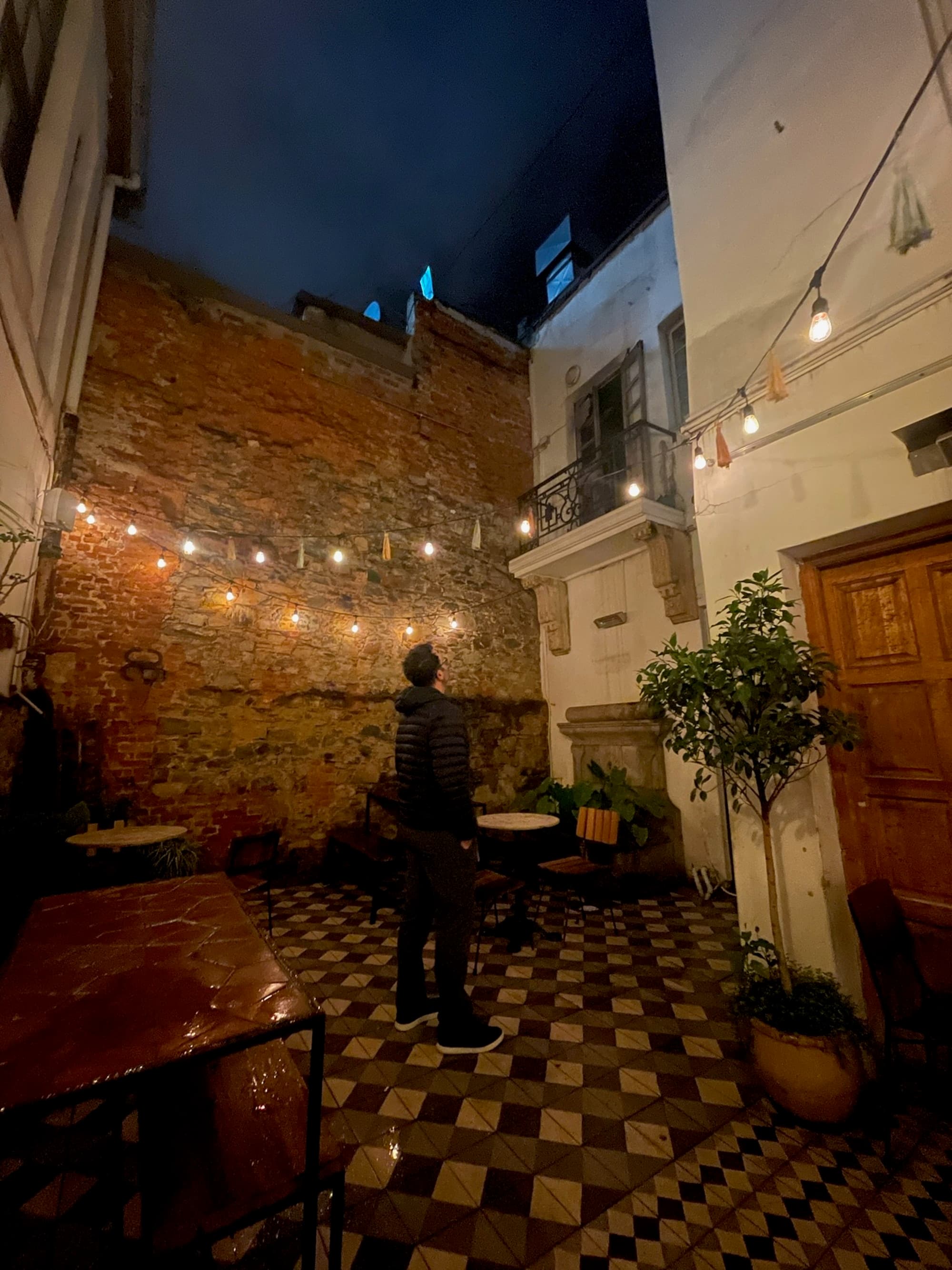 A person stands in a cozy, night-lit outdoor courtyard, surrounded by string lights and checkered tiles.