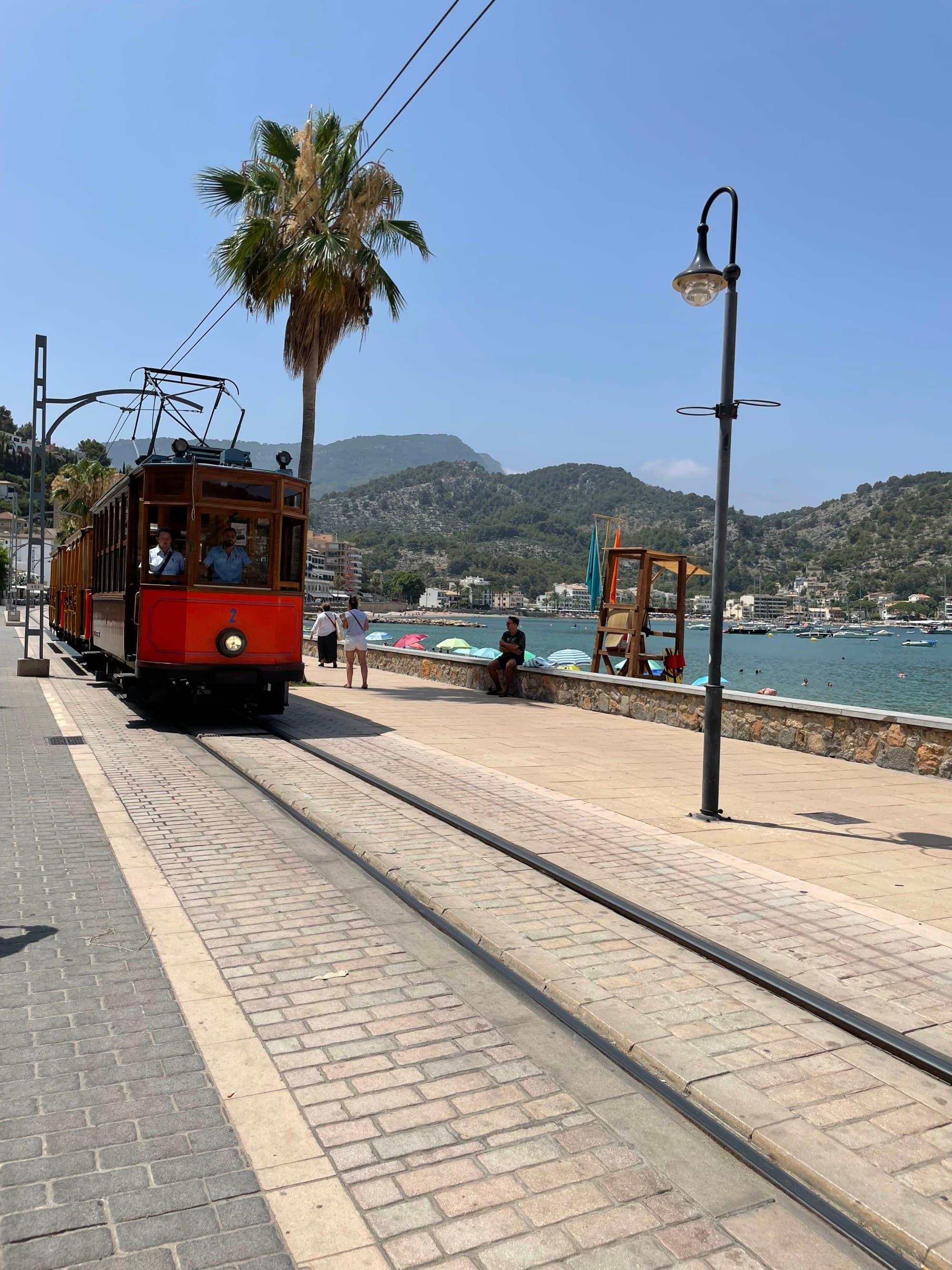 A red tram car chugging down a coastal road with the sea, mountains and blue sky in the background.