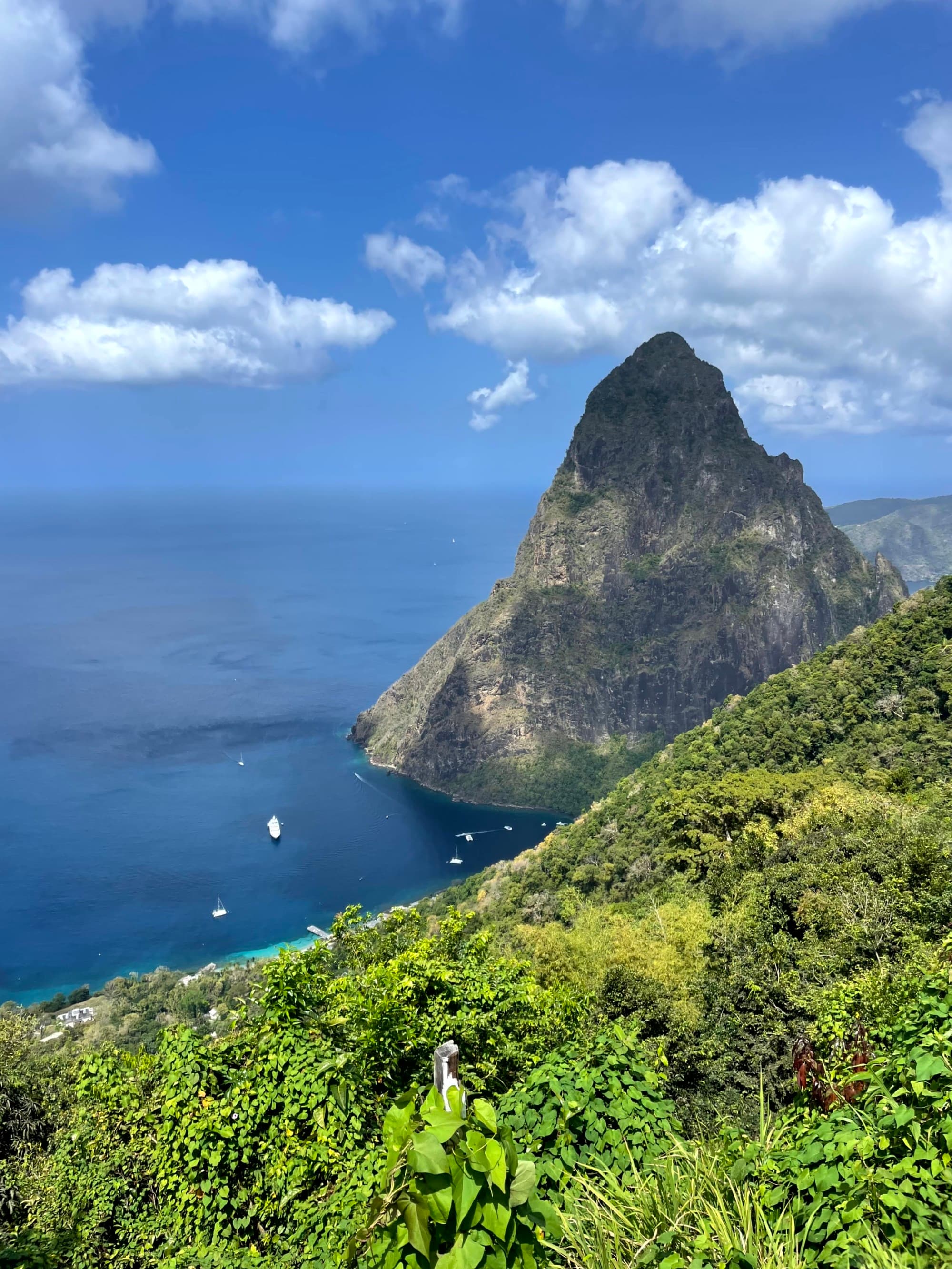 A picturesque green landscape with a mountain peak overlooking a blue bay dotted with boats under a partly cloudy sky.