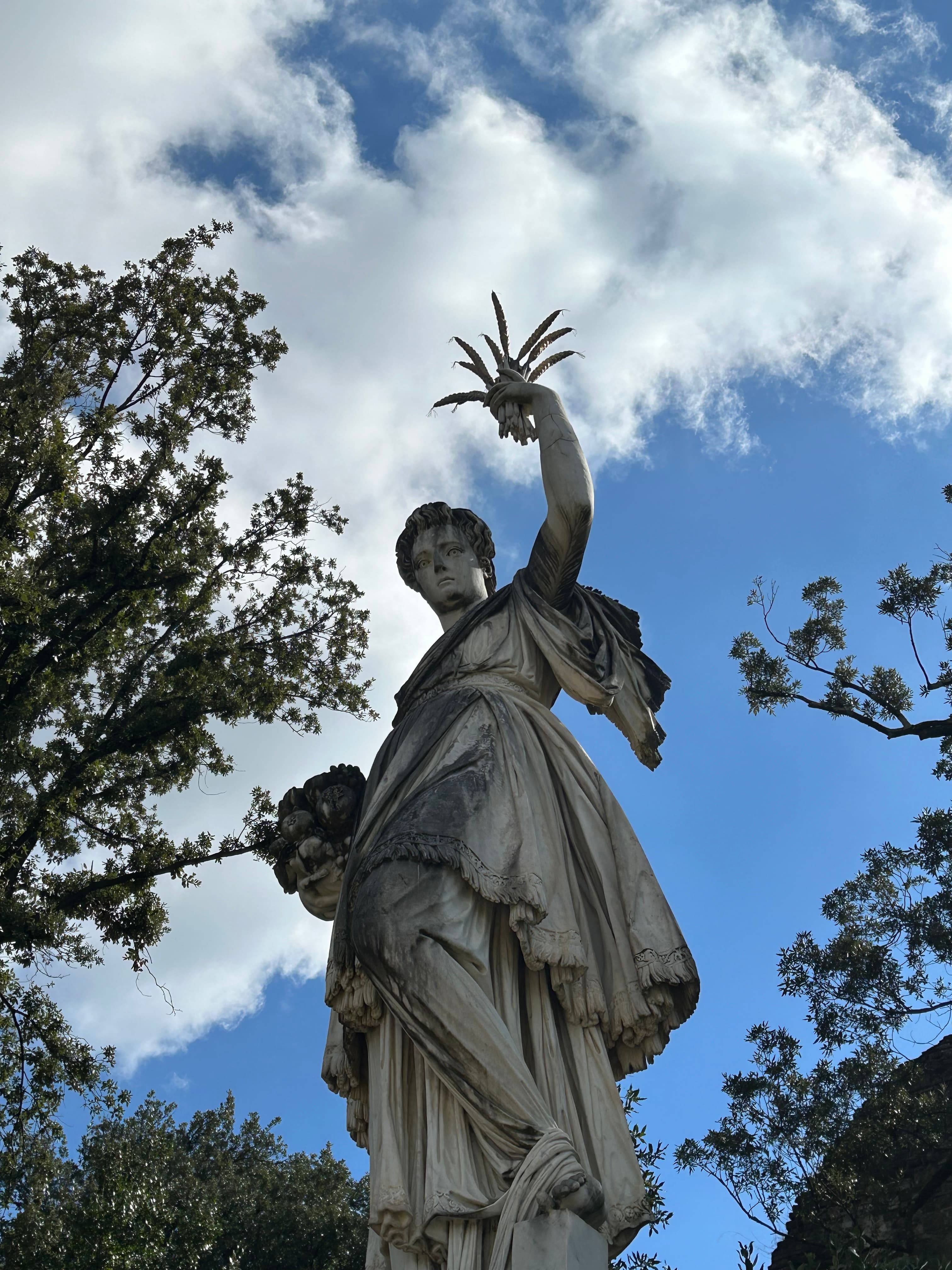 An ornate tatue with a cross, bathed in sunlight, towering over trees under clear blue skies.