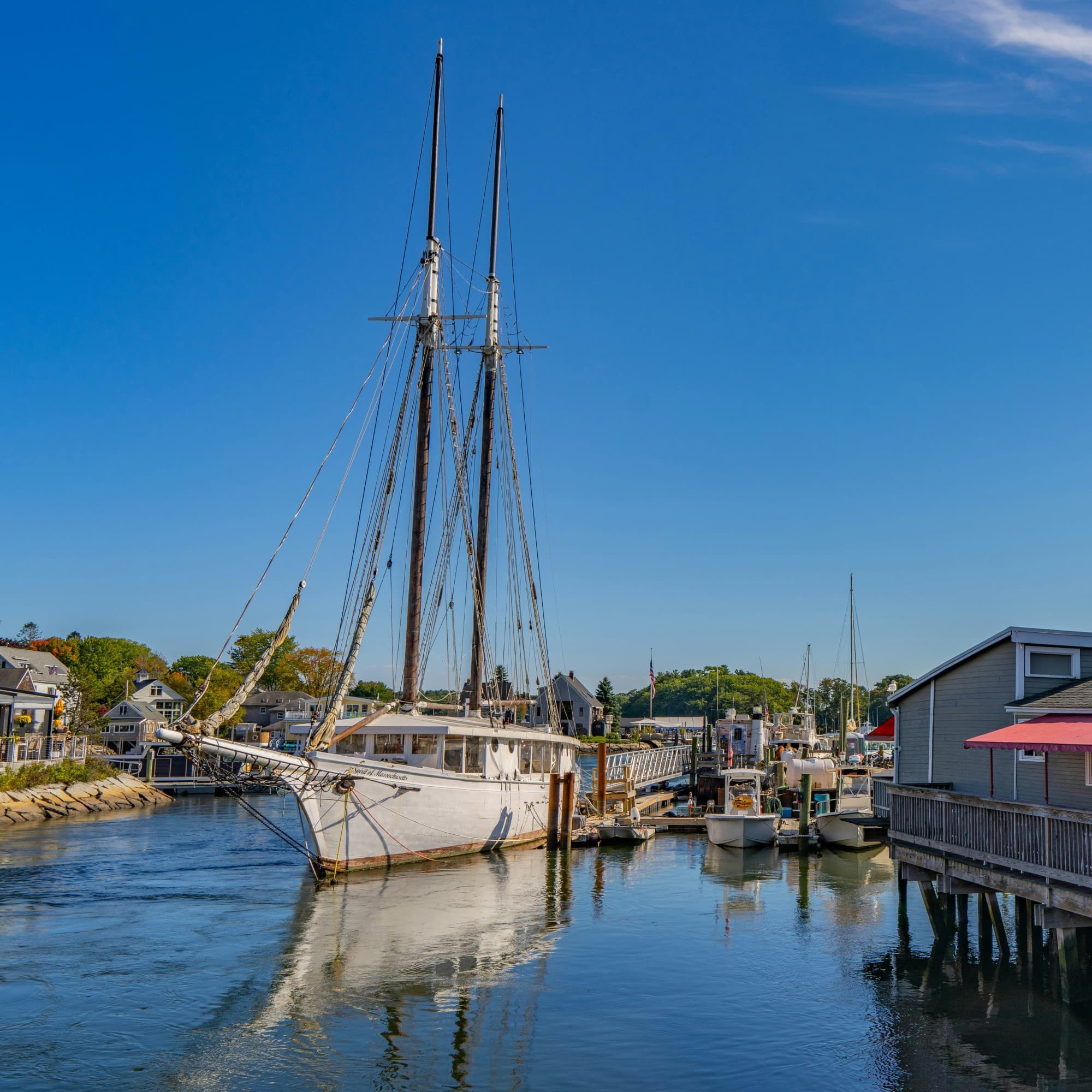 The image depicts a scenic harbor with a tall ship and “The Black Shack” waterfront building under a clear blue sky.