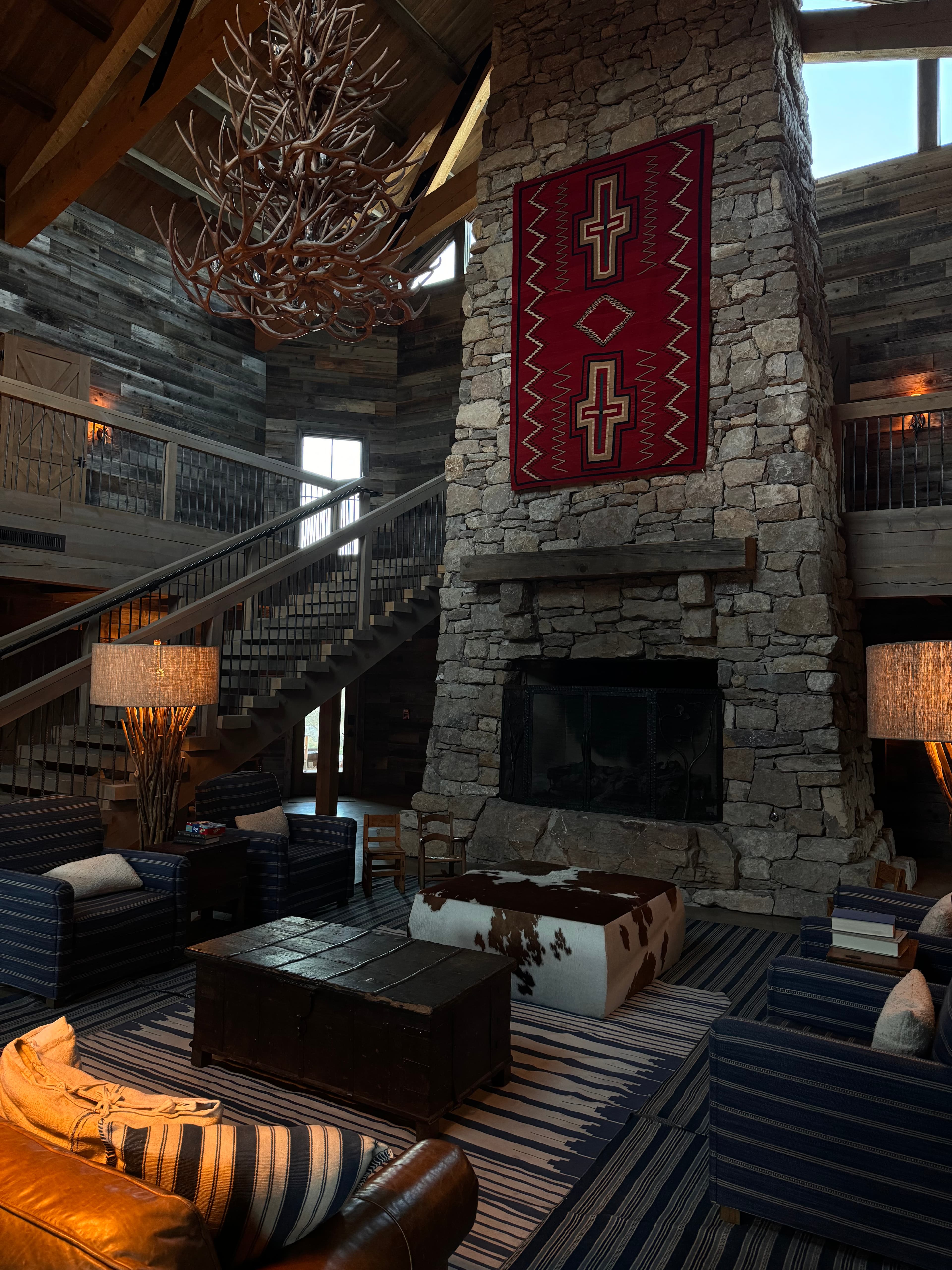 An image of a stone fireplace with a red carpet, wooden chandelier, seating area, leather couch, stairwell and carpet.
