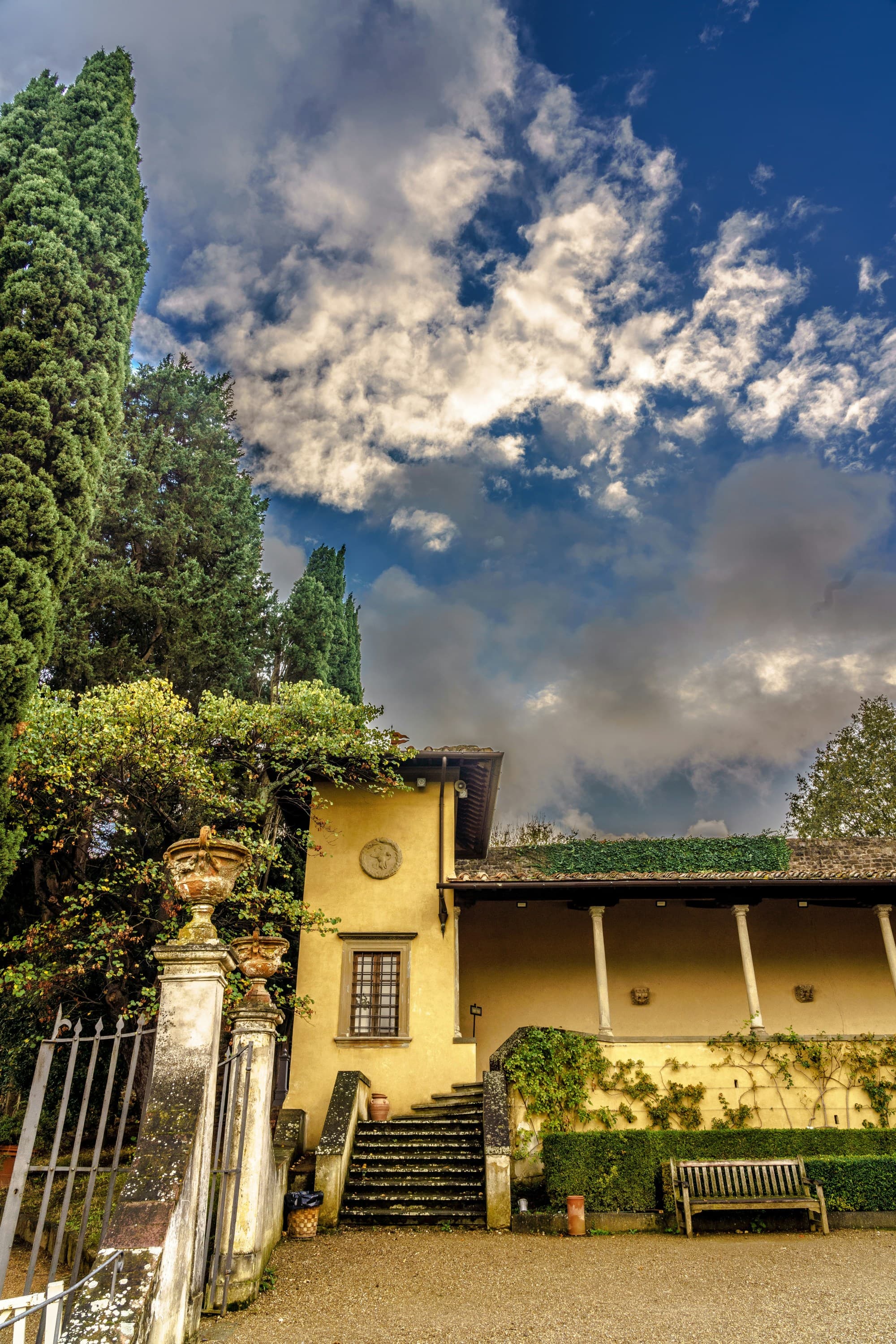 The image features a quaint yellow house with a staircase and lush greenery, under a sky with clouds, exuding old-world charm.