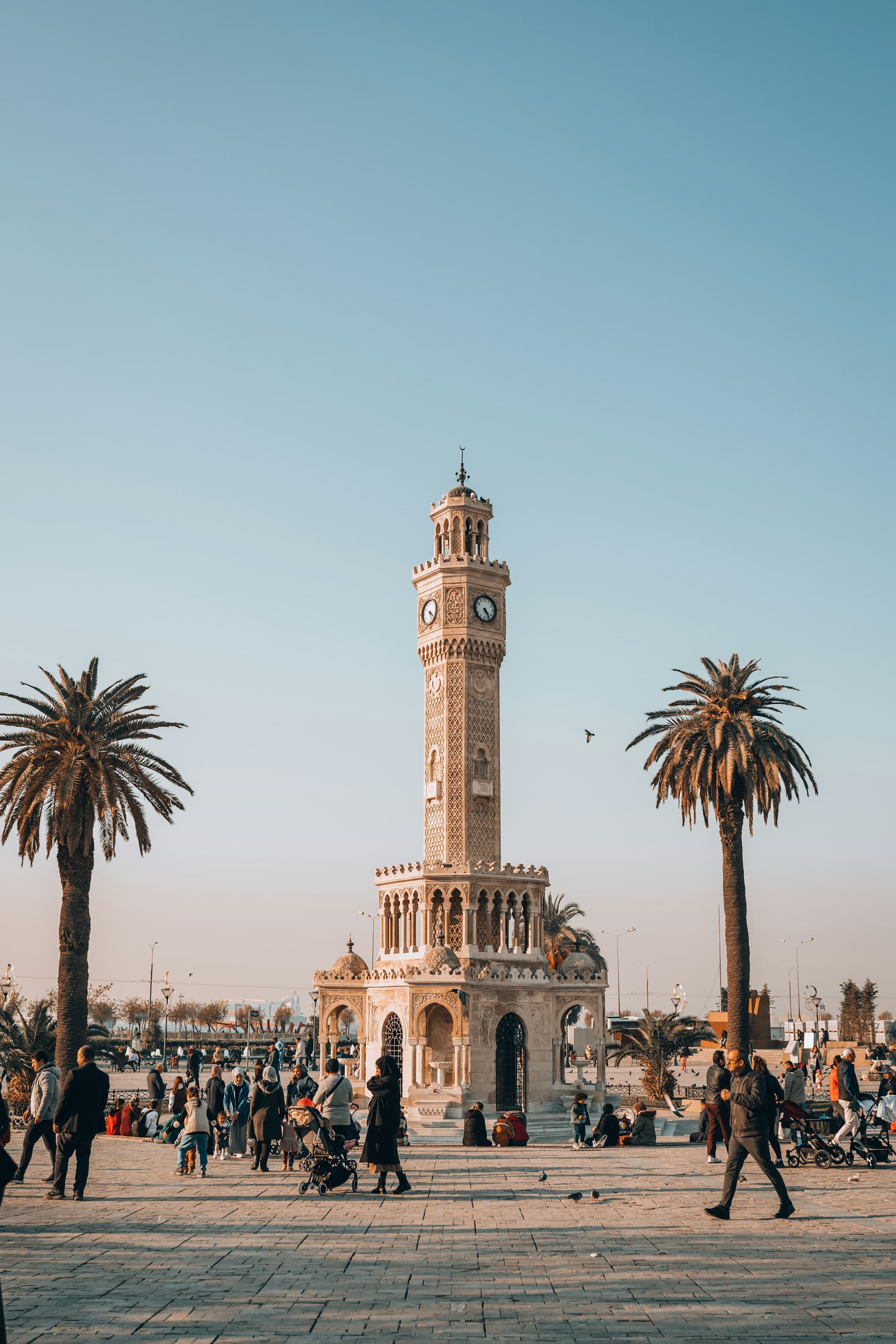 A tall, ornate clock tower in the middle of a busy square with many people walking around