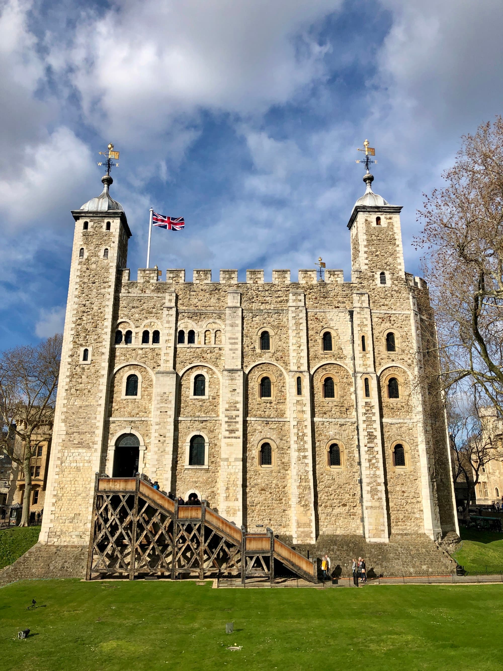 Old stone building with two turrets and a UK flag flying on the roof, with many small window openings and green grass in front