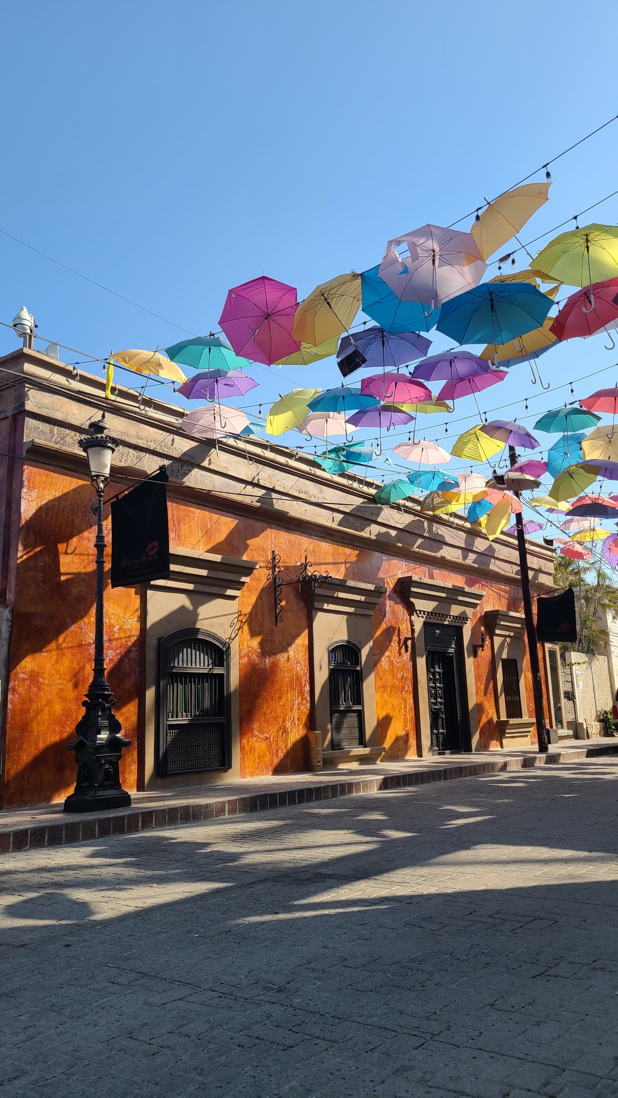 A street with brown walled buildings, post lights and colorful umbrellas above.