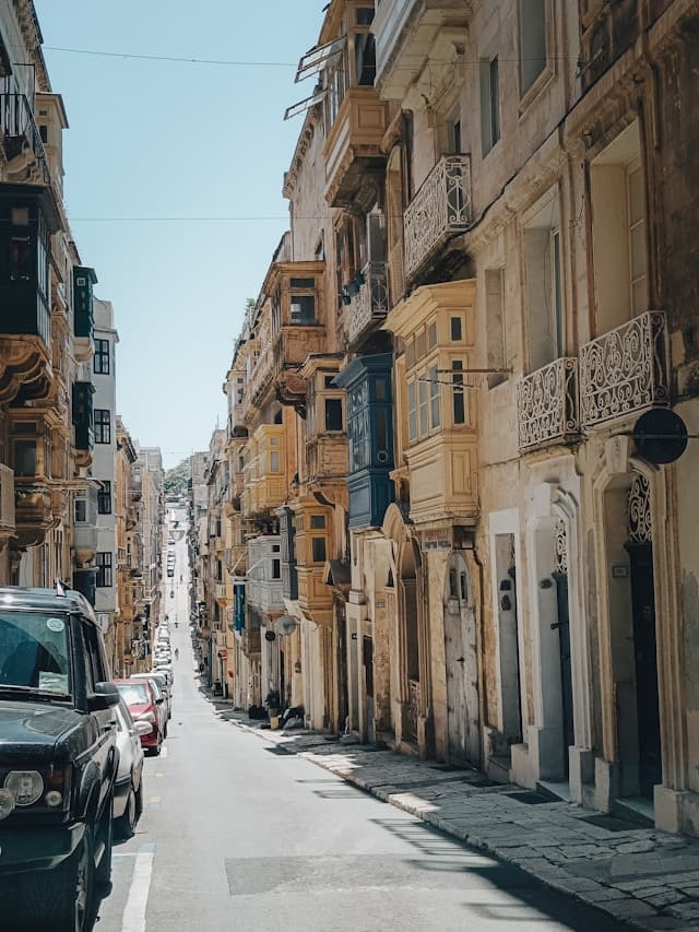 An image of a European city street with various stone balconies and detailing. There are cars parked along the left side of the street.